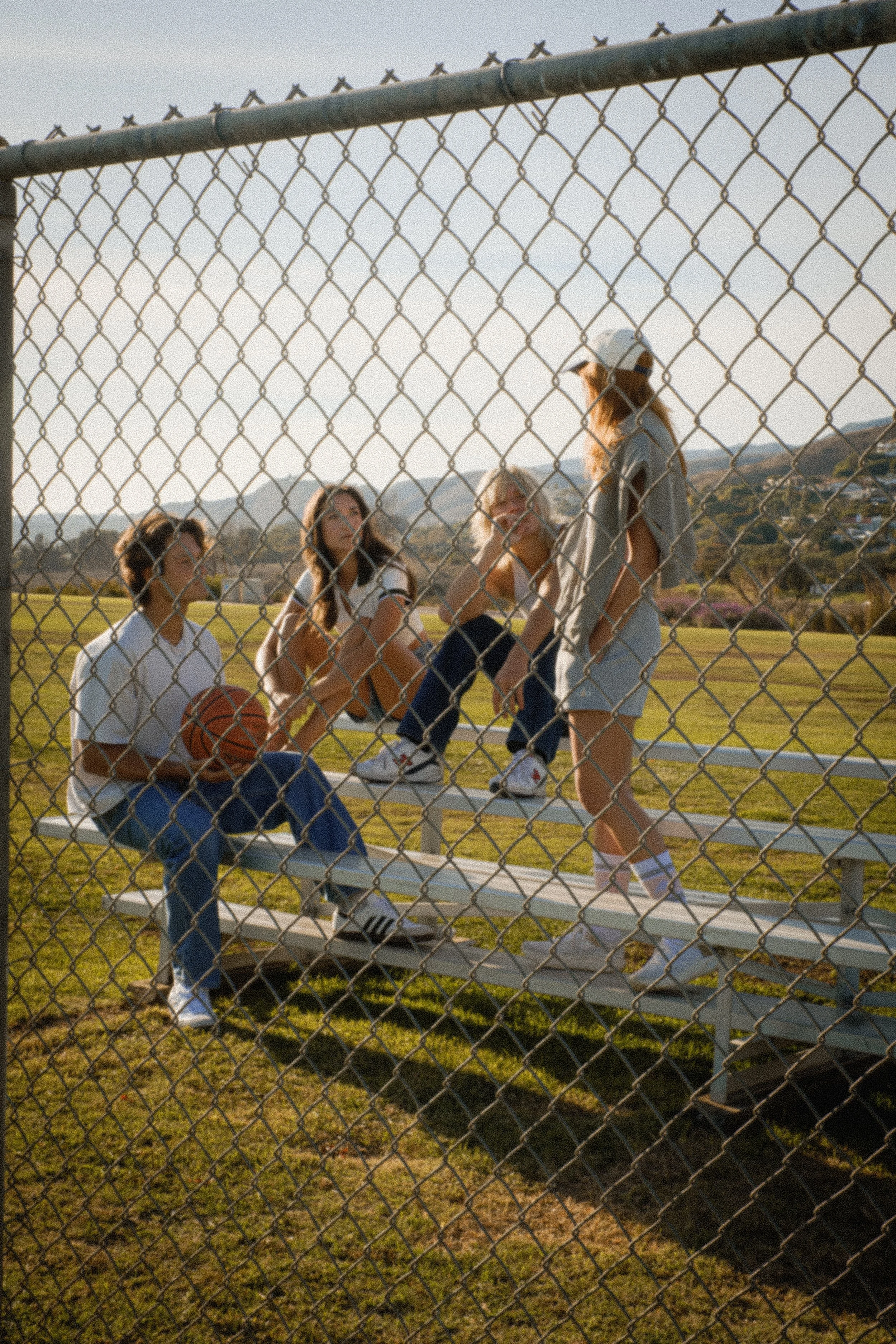 Four teenage girls are sitting on bleachers at a baseball field, with one girl standing and talking to the others. They are behind a chain-link fence, with mountains in the background. The scene is during late afternoon or early evening.