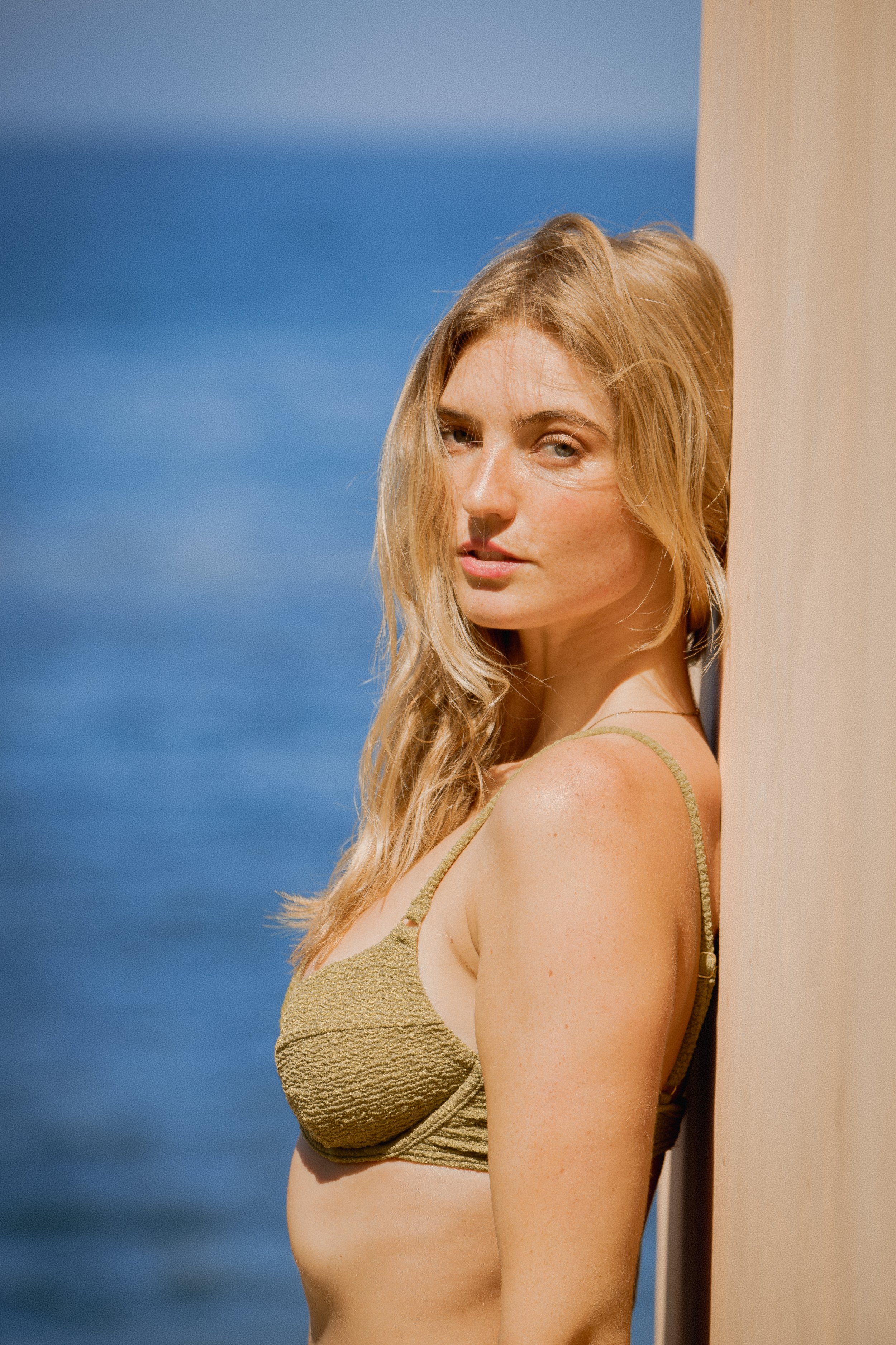 A woman with long blonde hair leaning against a beige column outdoors with a blue sky background.