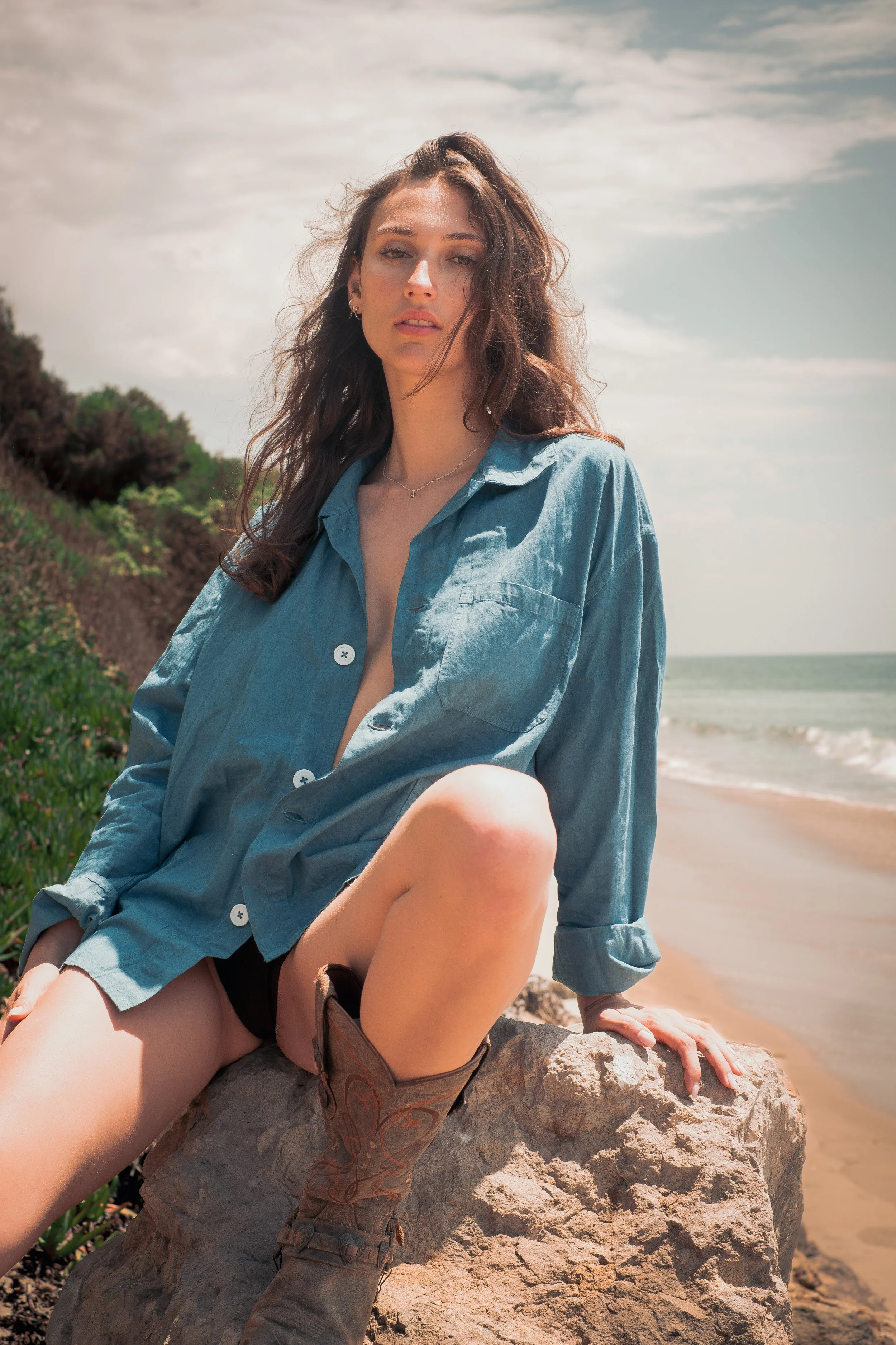 A young woman sitting on a large rock on a beach with the ocean and cloudy sky in the background. She has long, wavy brown hair, is wearing a blue shirt, black shorts, and cowboy boots, and gazes at the camera.