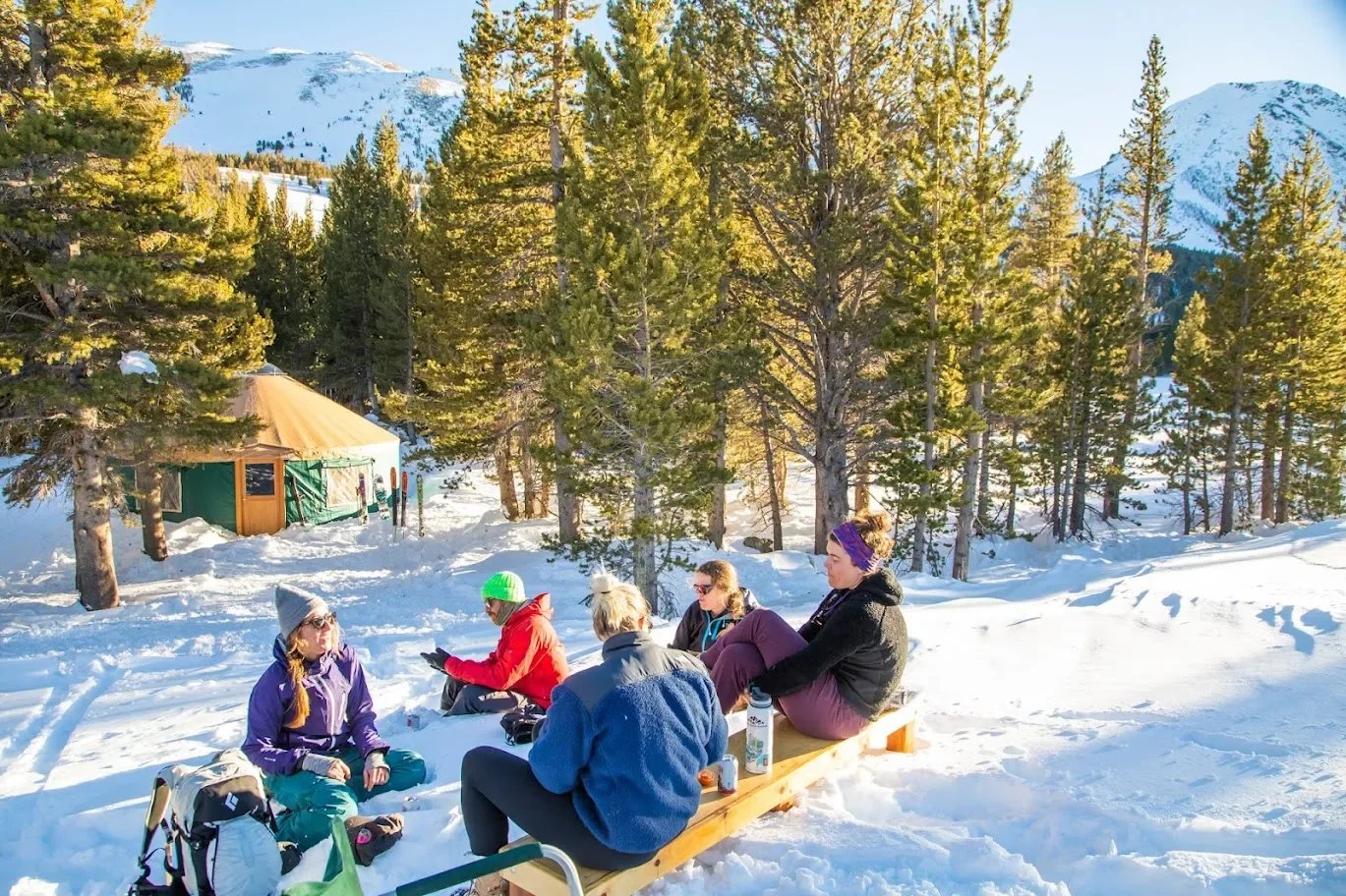 Group of people sitting on a wooden bench in a snowy forest with mountains in the background, enjoying a sunny winter day.