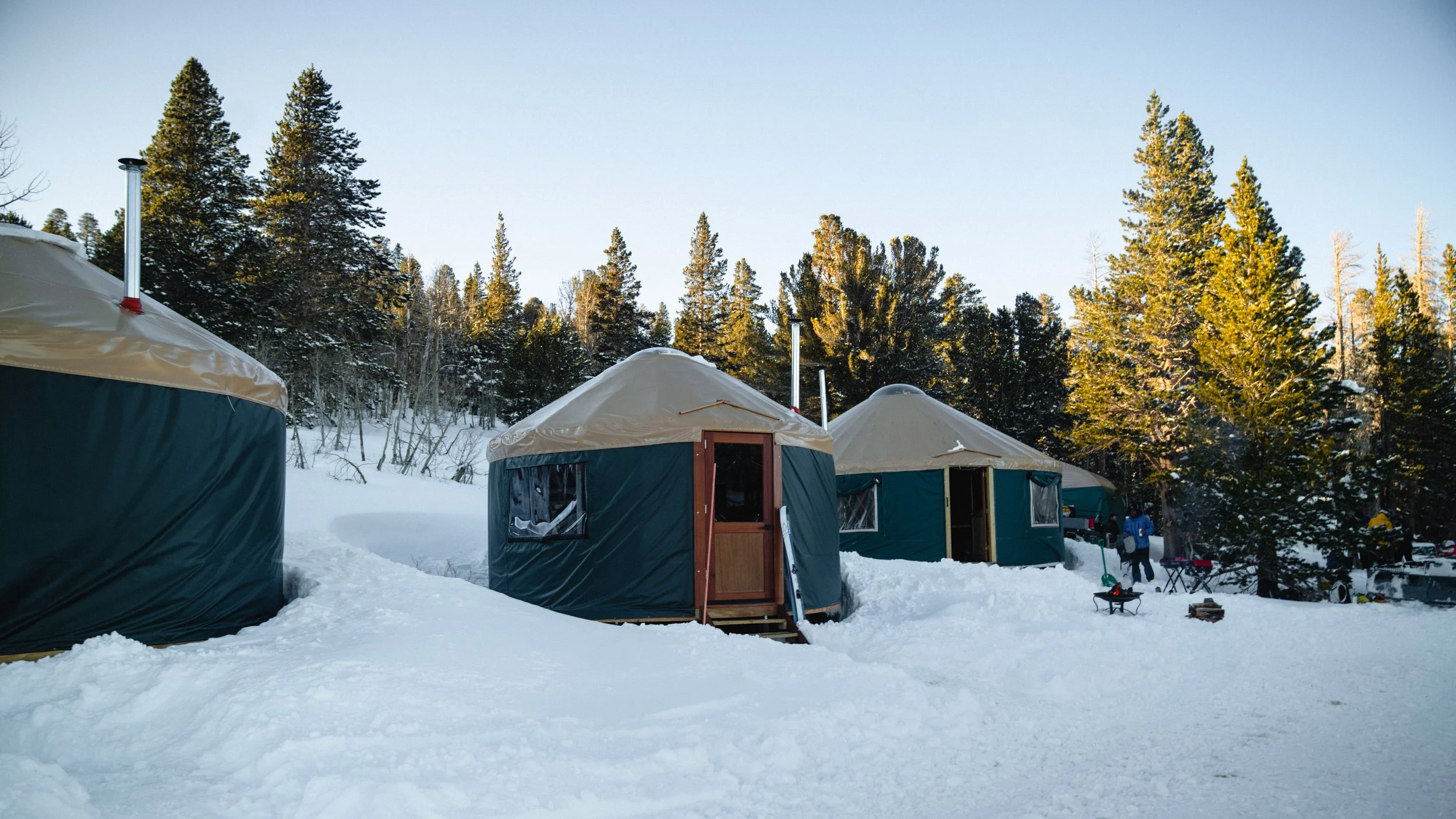A winter scene at a snowy campsite with multiple yurts and people in the background, surrounded by tall pine trees and clear blue sky.