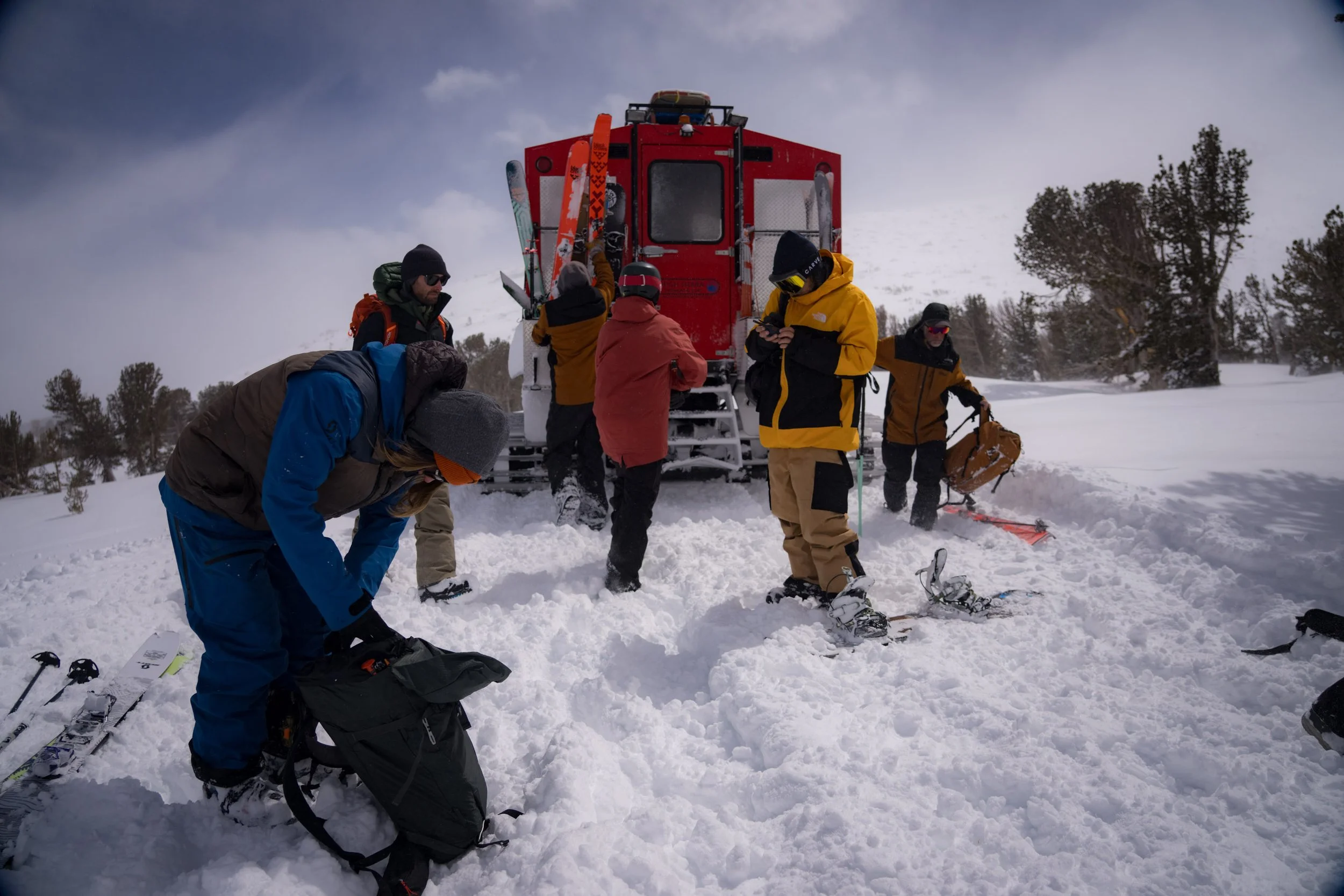 Group of people dressed in winter gear standing around a red snowcat in a snowy landscape with trees in the background.