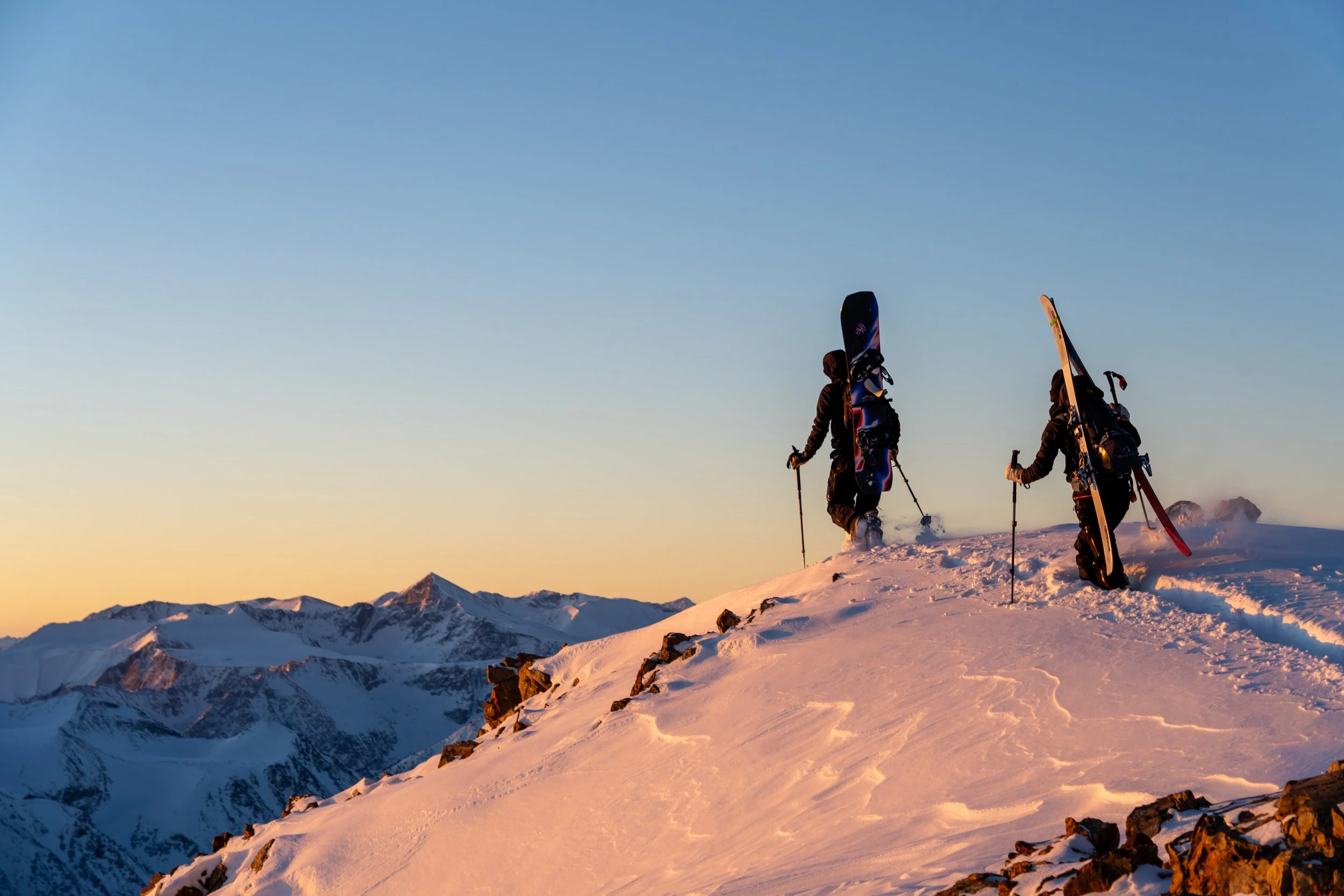 Two skiers carrying skis on their shoulders standing on snowy mountain ridge at sunset with mountain range in background.