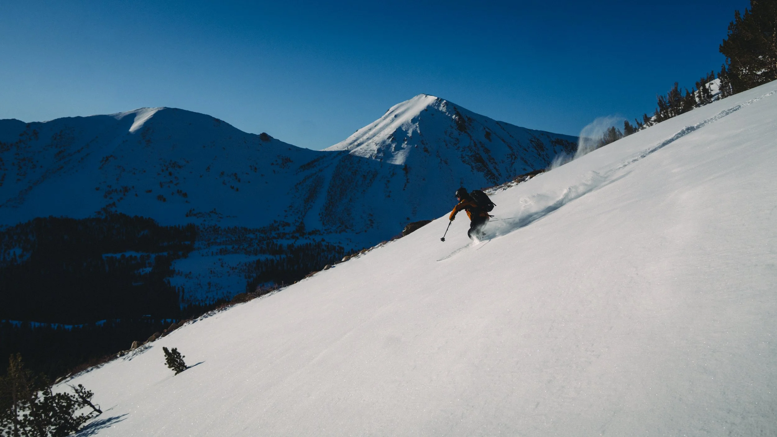 A person snowboarding down a snowy mountain slope in a mountainous landscape with snow-covered peaks and partly cloudy sky.