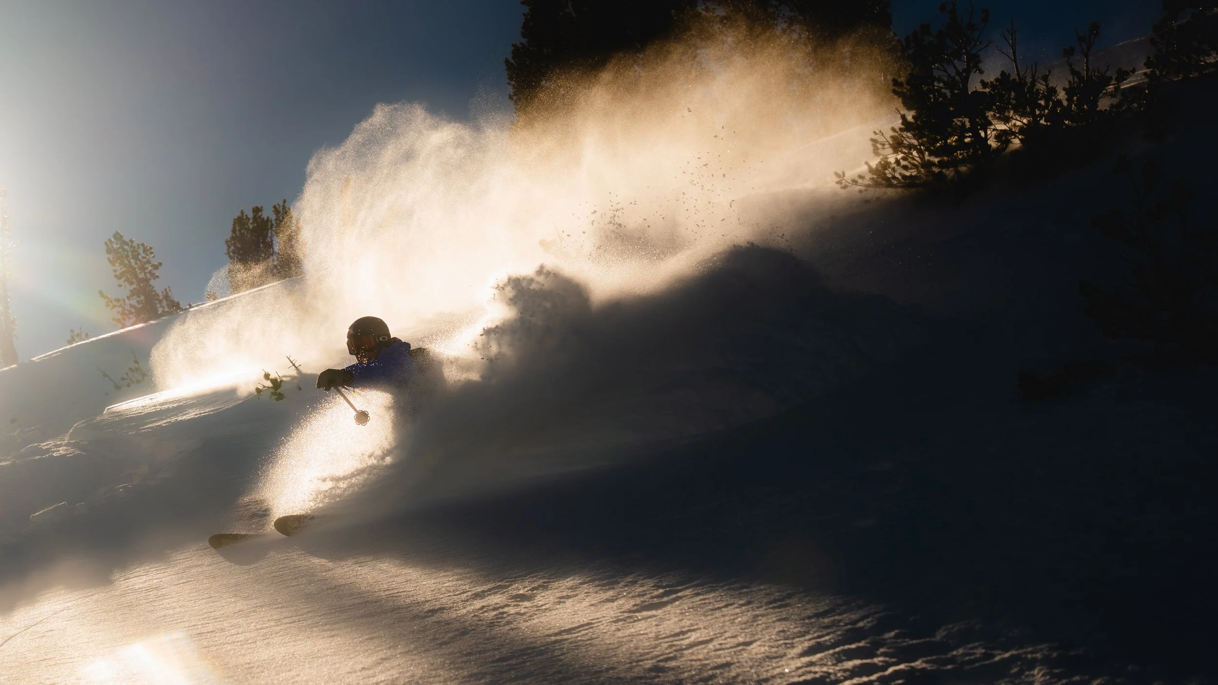A skier wearing a helmet speeds through fresh powder snow, creating a large spray of snow behind them in a forested mountain area during sunset.