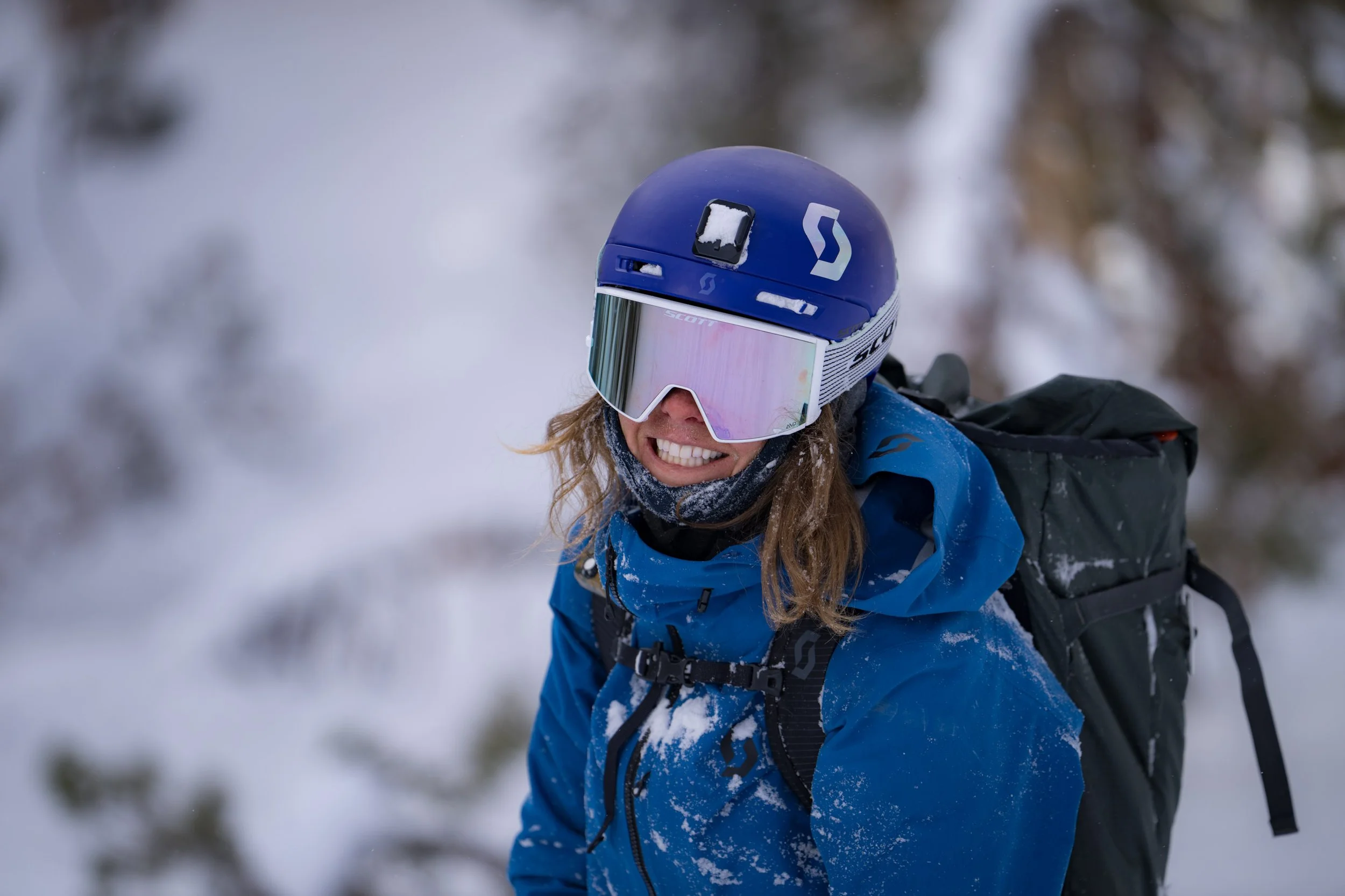 A woman in winter gear, including a blue jacket, black backpack, helmet, and ski goggles, is smiling and skiing through snow with a blurred snowy forest background.