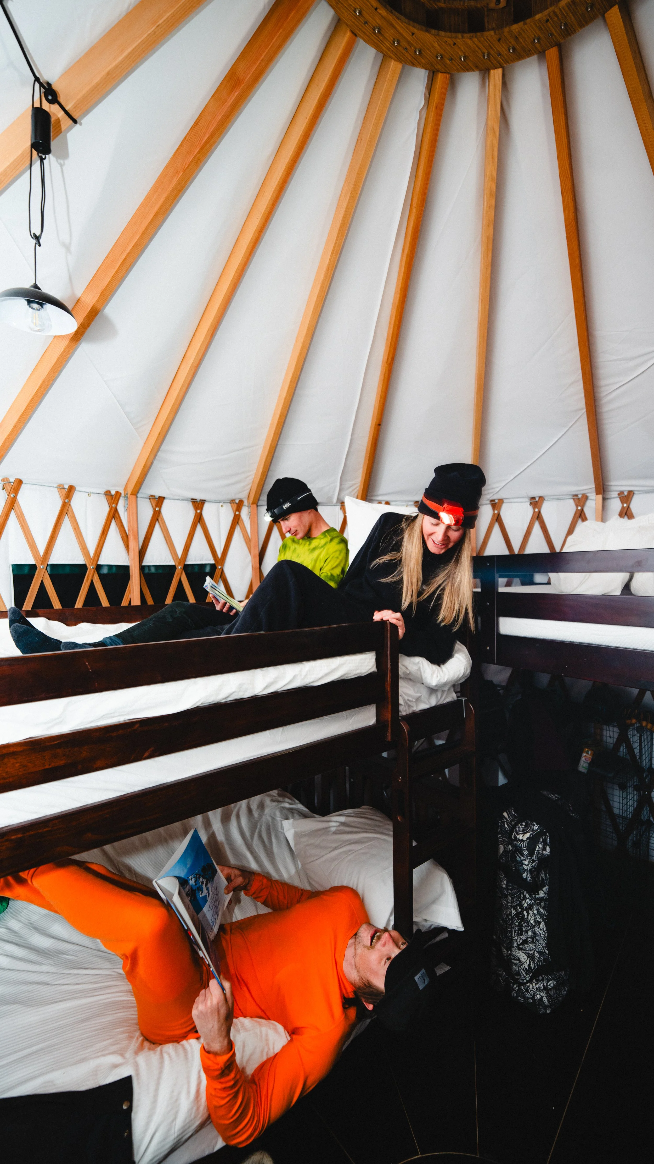 Interior of a yurt with people lying on beds, one using a laptop, others reading or using a smartphone, with wooden beams and a fabric wall.