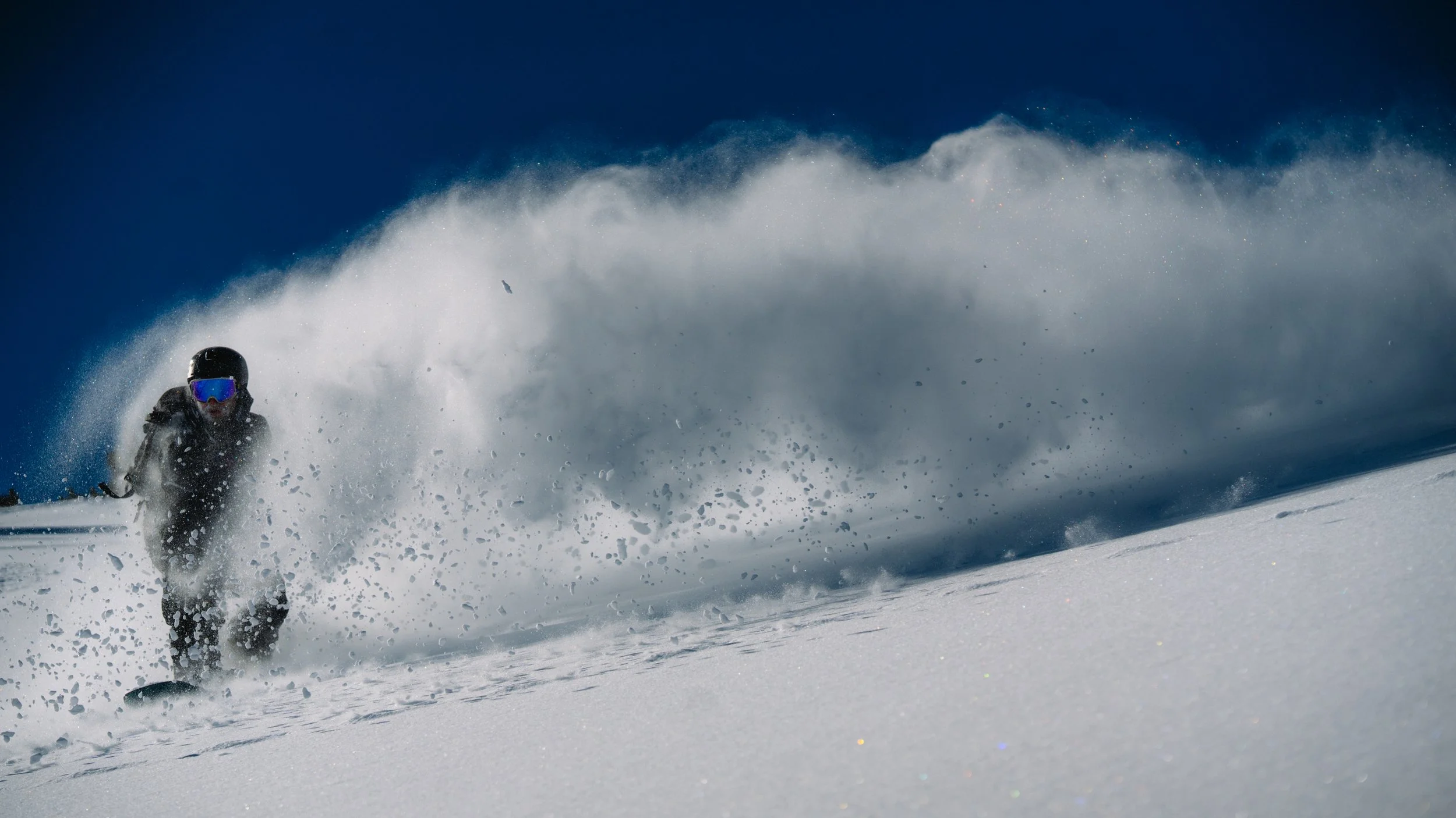 A person skiing down a snowy mountain slope, surrounded by a spray of snow and a clear blue sky.