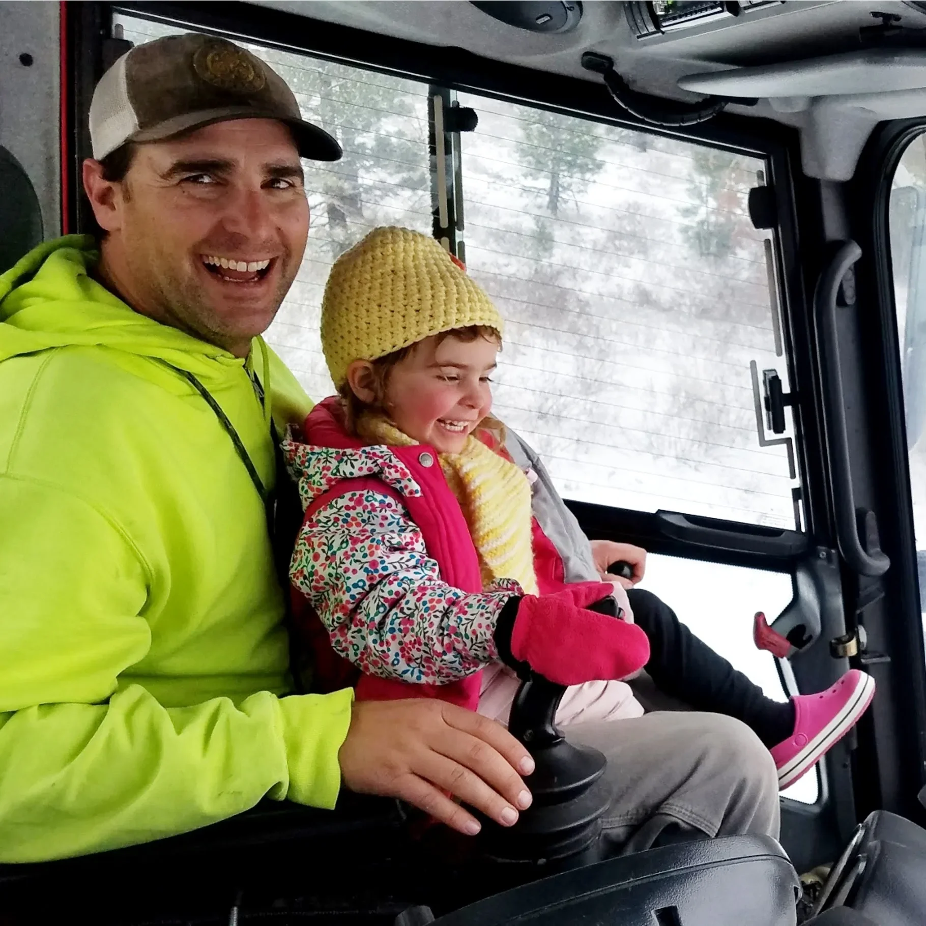 A smiling man wearing a high-visibility yellow jacket sitting next to a young girl in a vehicle. The girl is wearing a yellow knit hat, a colorful patterned jacket, and pink rain boots. They are inside a vehicle with a windshield showing a snowy landscape outside.