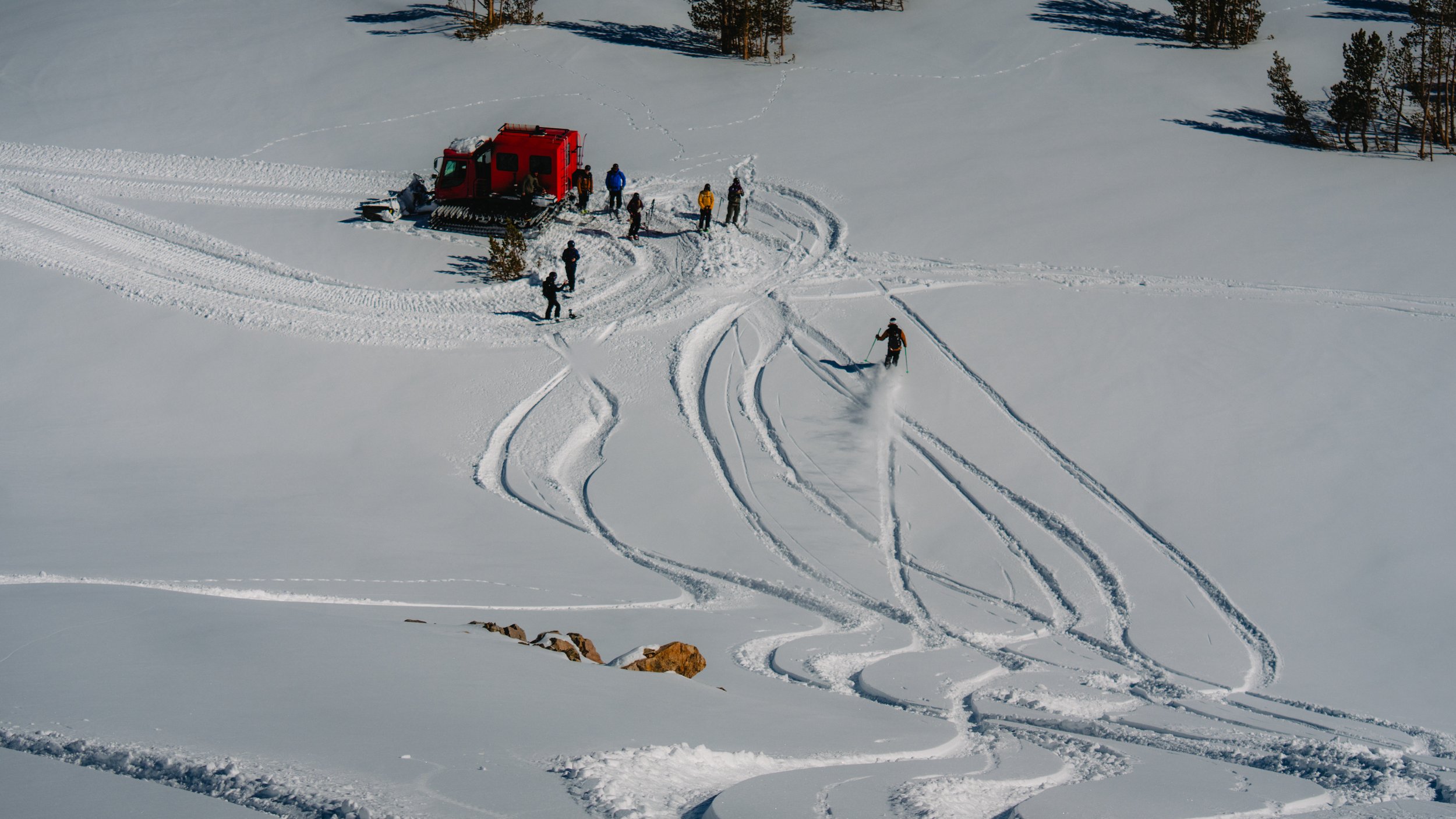 A group of people snowmobiling in a snow-covered landscape with trees in the background and a red snowcat vehicle nearby.