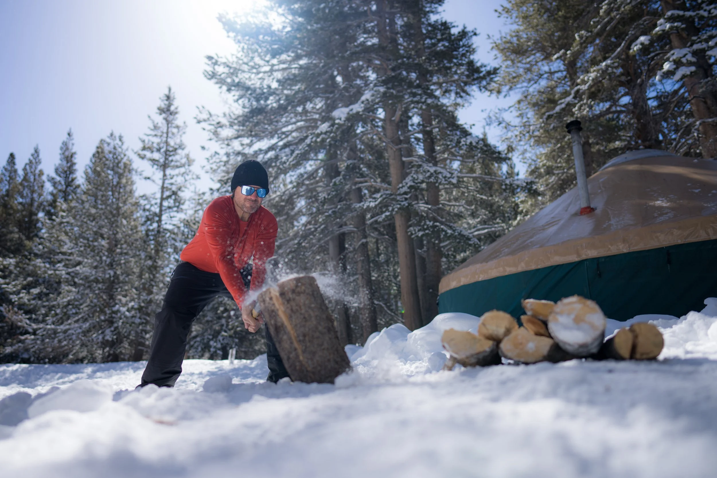 Man chopping wood in a snowy forested area near a tent.