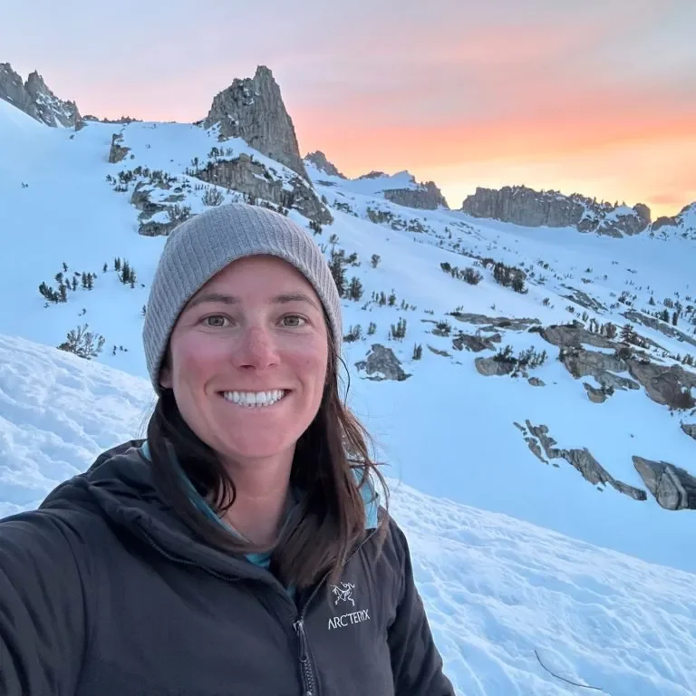 A woman taking a selfie in a snowy mountain landscape during sunset, wearing a gray beanie and black Arc'teryx jacket.