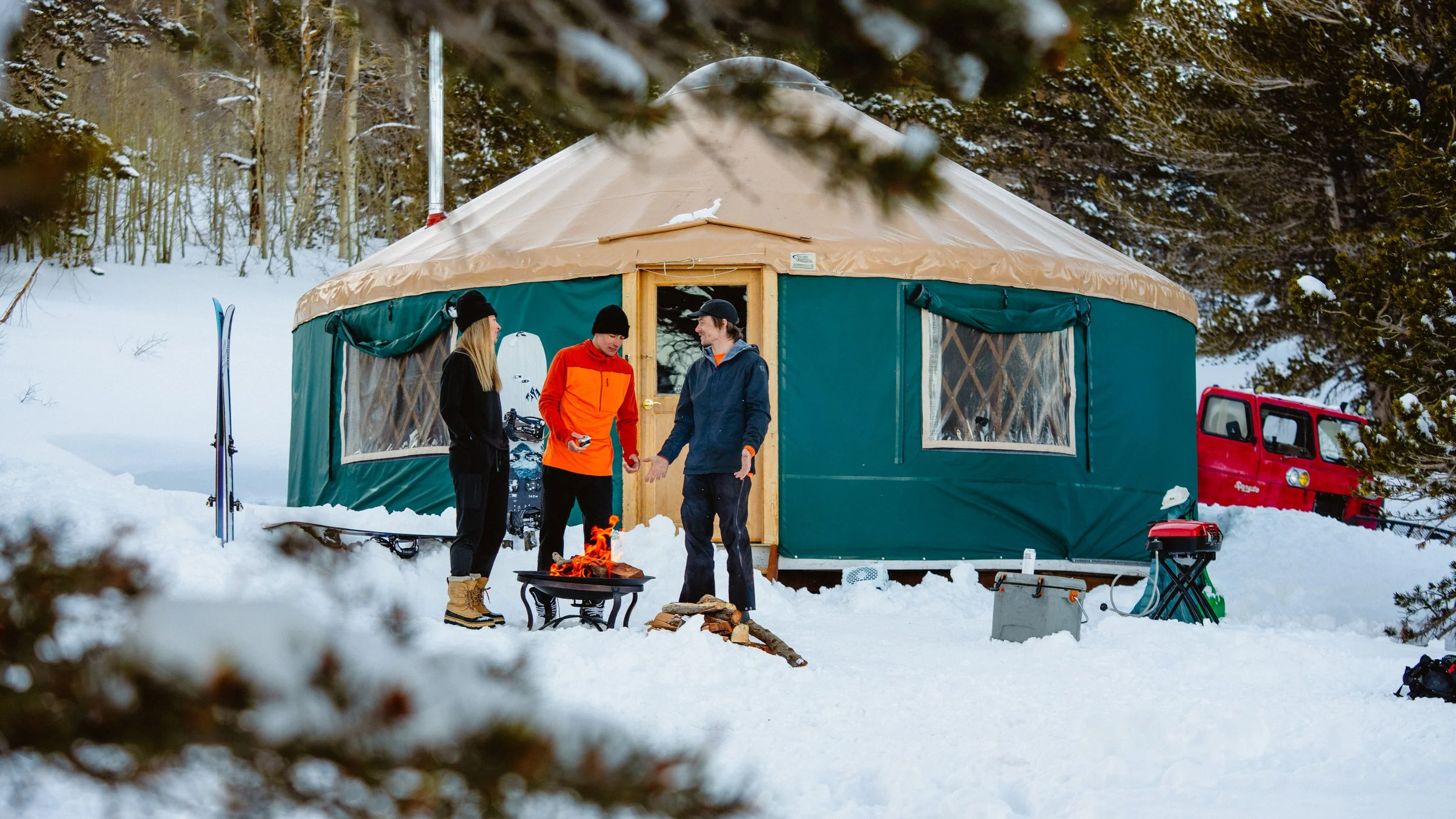 Three people standing outside of a large tent in a snowy forest, talking near a small campfire, with skis and a red vehicle nearby.