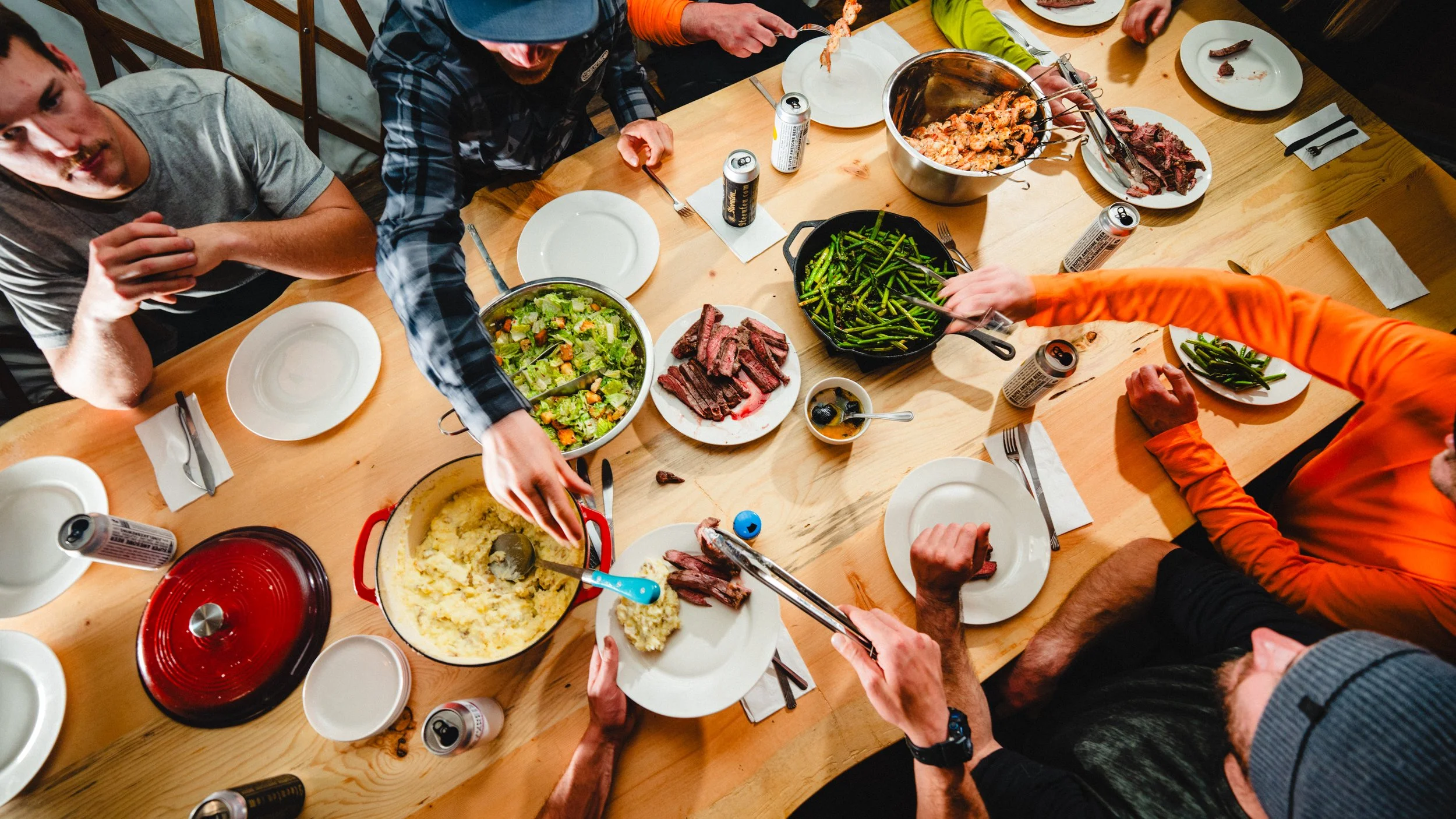 People gathered around a wooden table enjoying a meal with various dishes including mashed potatoes, green beans, salad, sliced steak, and shrimp, along with cans of soda.