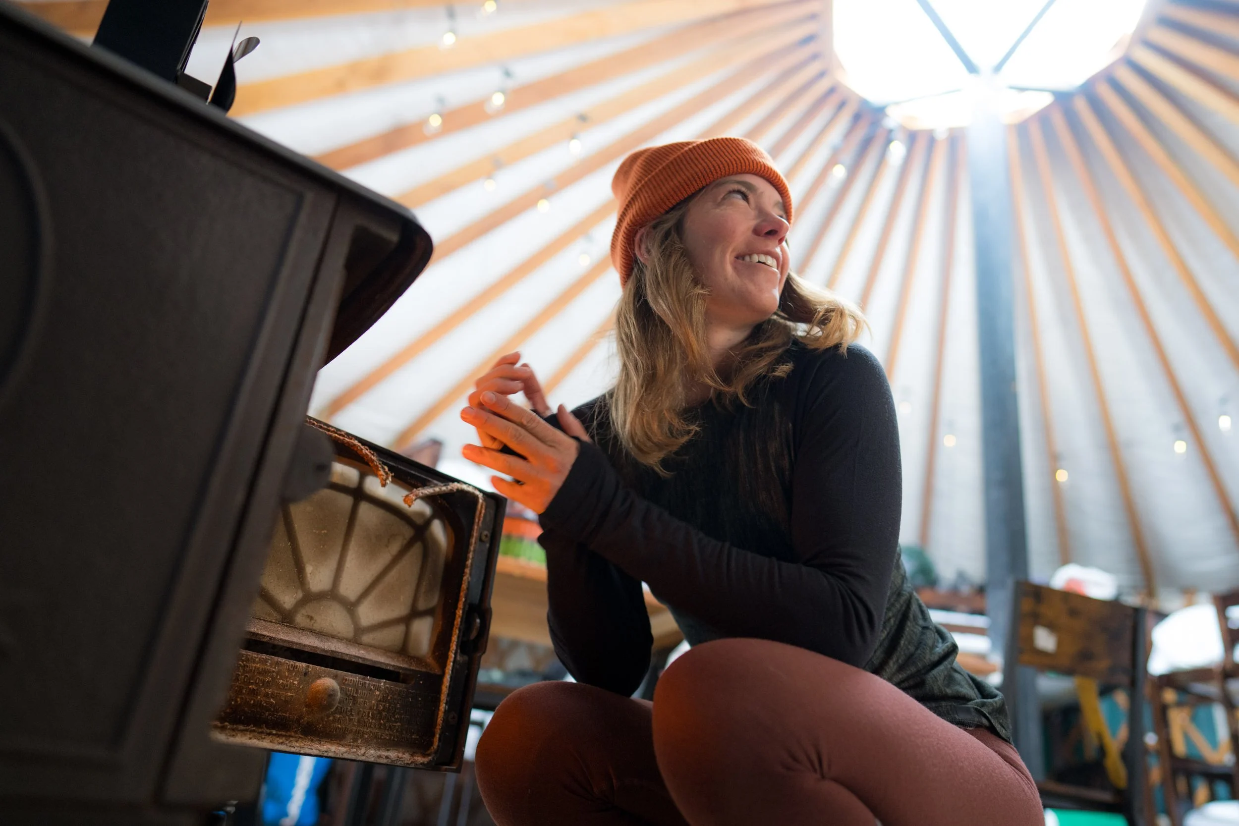 A woman wearing an orange beanie and black jacket sitting near a piano in a wooden-ceiling room with sunlight coming through the roof.