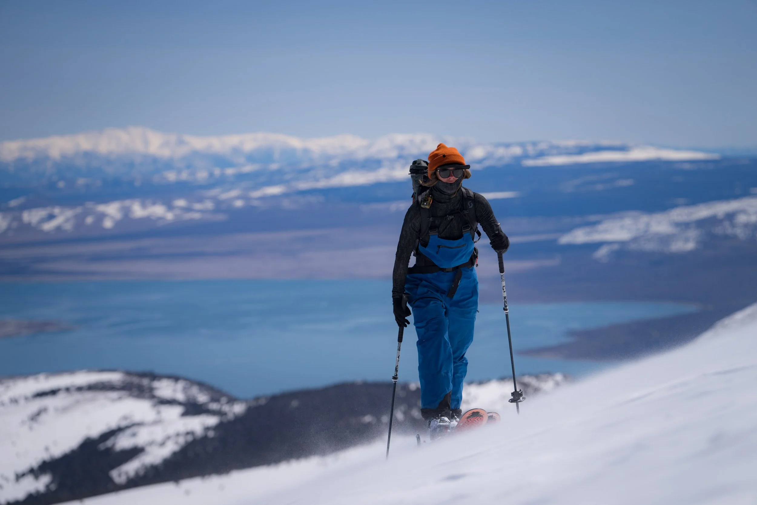A person in outdoor winter clothing, including an orange hat, sunglasses, and face covering, skiing on snow-covered terrain with a mountain range in the background.