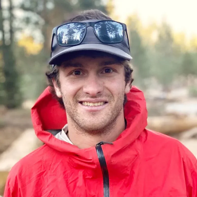 Man outdoors wearing a red jacket, black cap, and sunglasses on top, smiling at the camera with trees in the background.