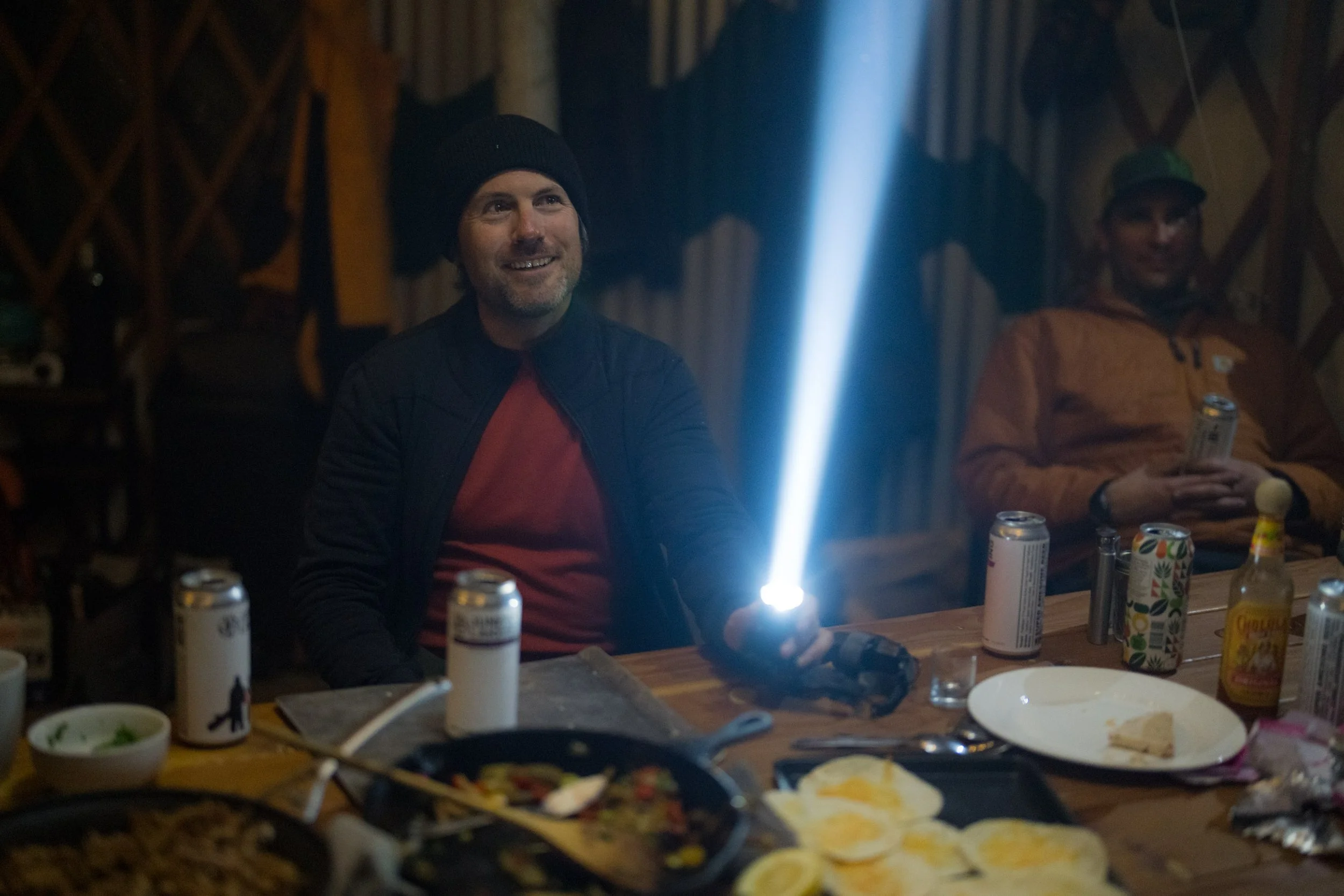 A man with dark hair and a beard smiling, wearing a black beanie and a black jacket over a red shirt, sitting at a dinner table in a wooden room, illuminated by a chat light.