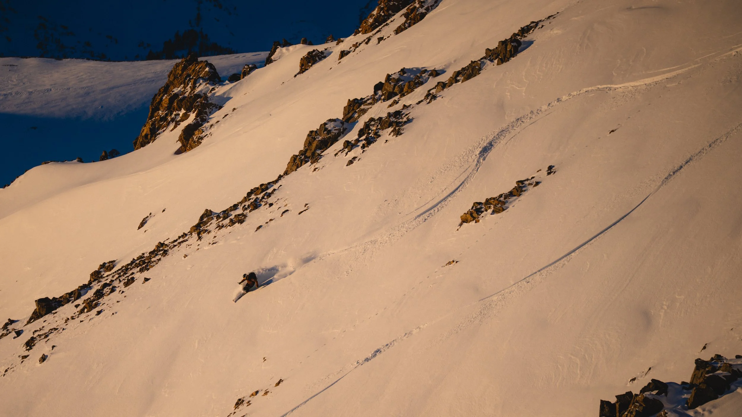 A skier descending a snowy mountain with rocky outcroppings, captured during sunset or sunrise.
