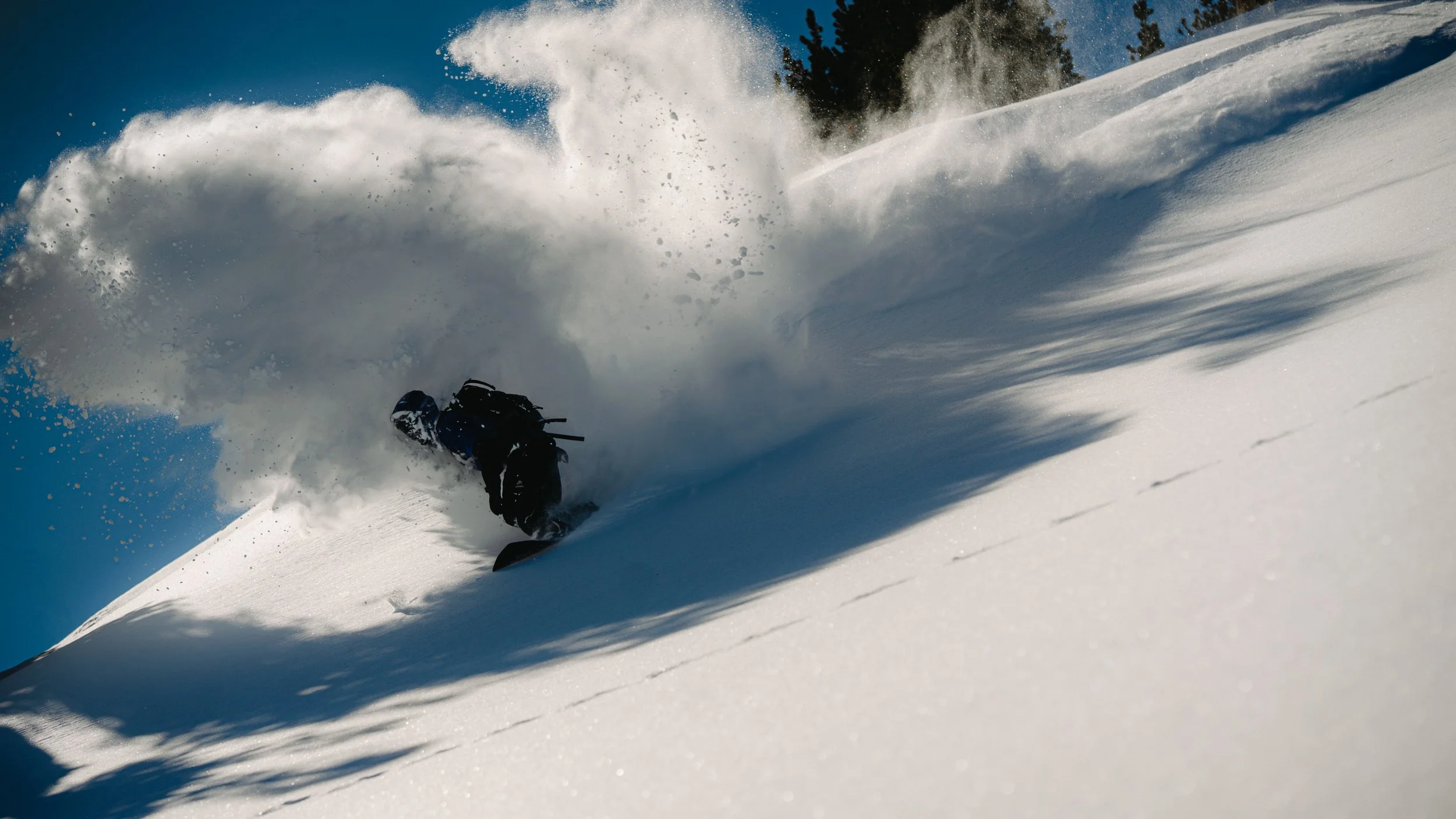 Person snowboarding down a steep snowy slope, kicking up a large spray of snow under a clear blue sky, with evergreen trees visible in the background.
