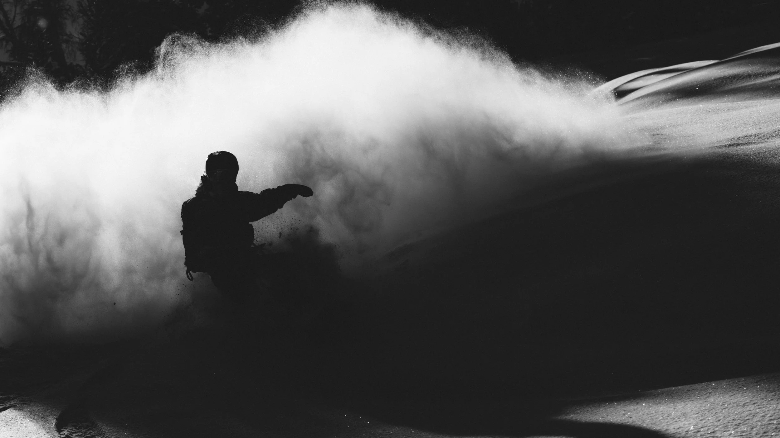 A person snowboarding on a snowy slope during daytime, with snow spray in the air and blurred trees in the background.