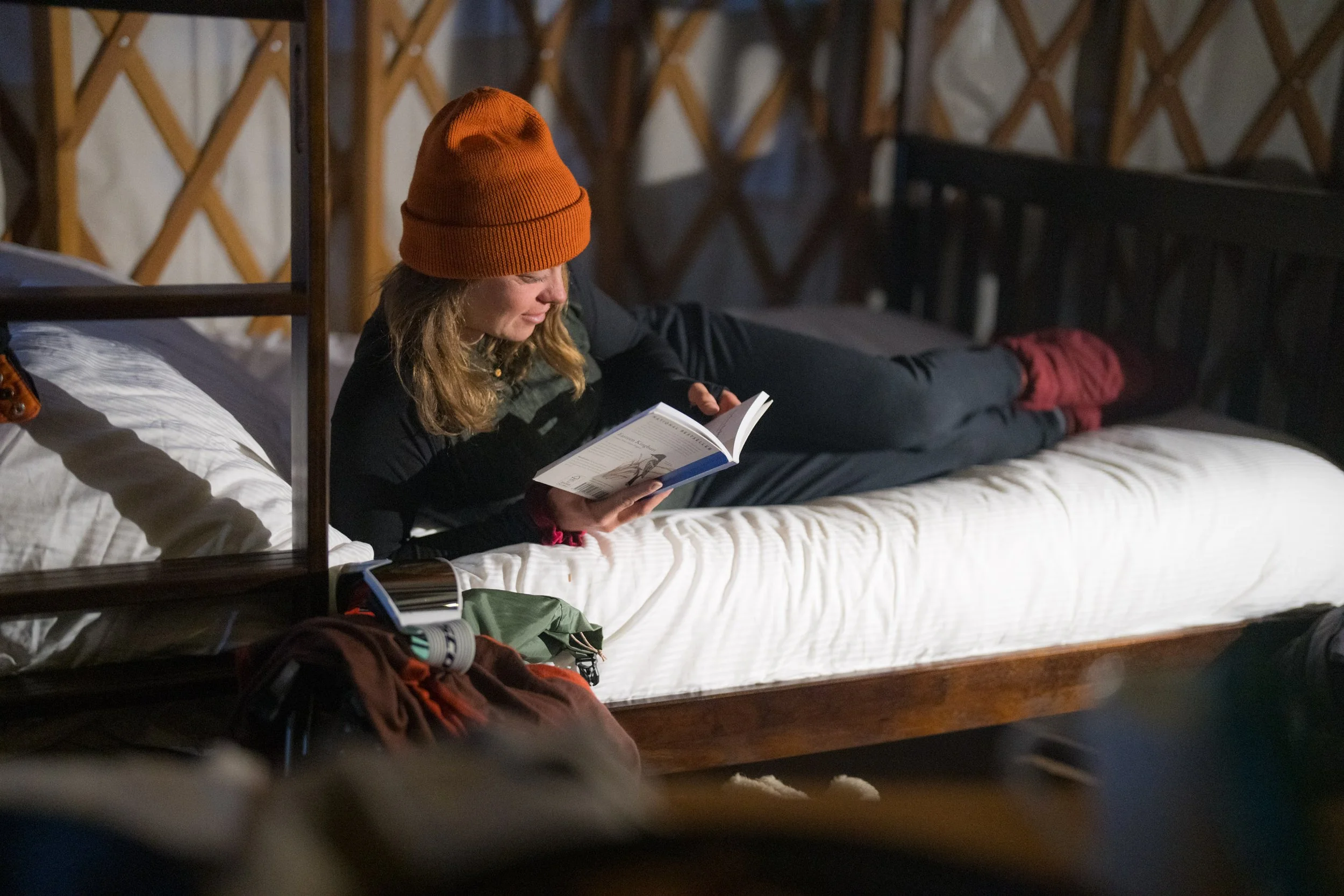 A woman with long blonde hair, wearing an orange knit beanie, black jacket, is lying on her stomach on a bed, reading a book in a cozy, warmly lit room with a wooden bedframe and quilted blanket.