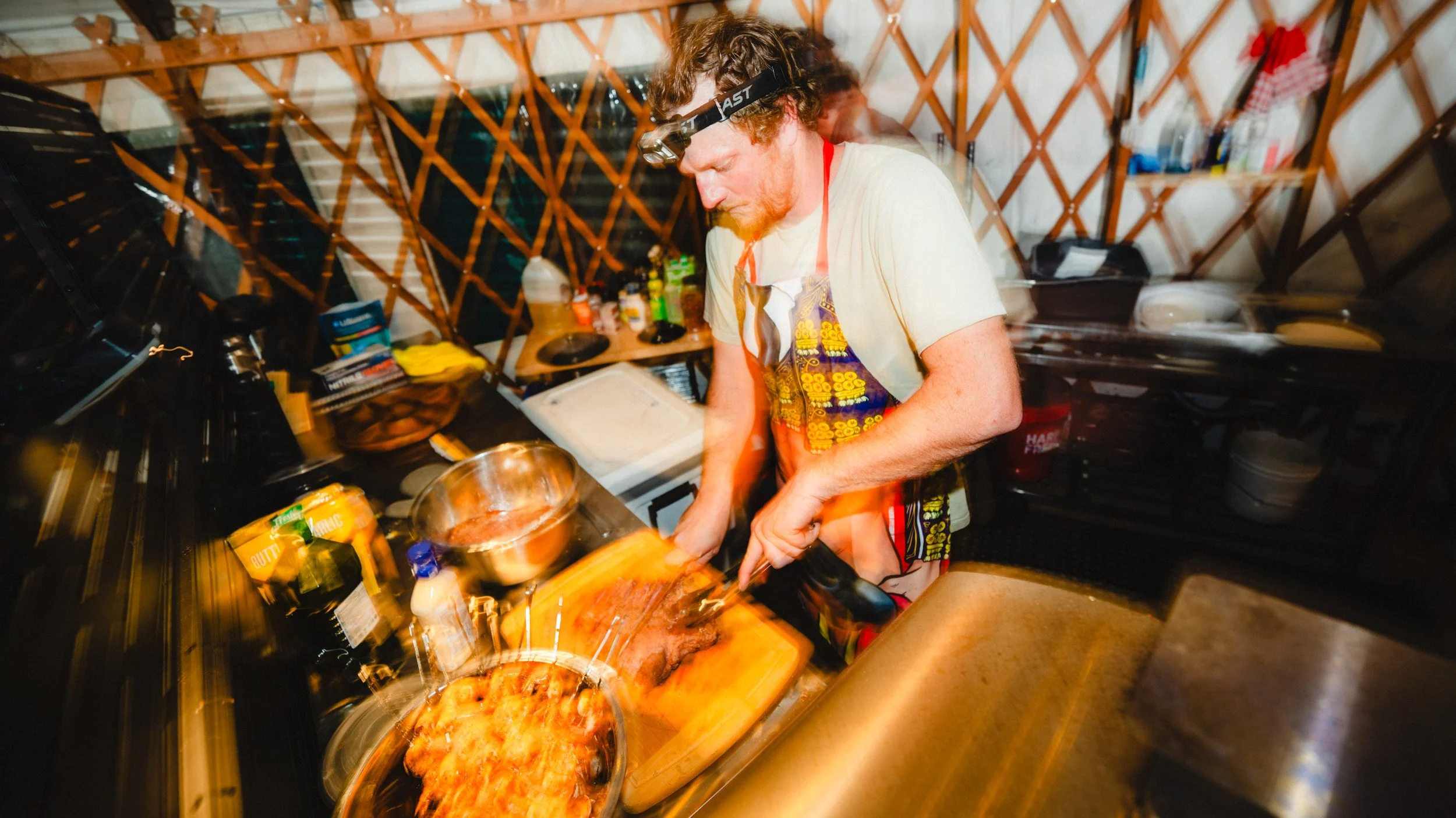 A man with red hair and a beard, wearing a headlamp and apron, is preparing food in a yurt kitchen, chopping meat with a knife.