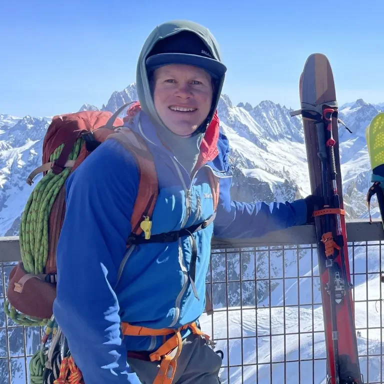 A person in winter gear standing in the mountains with snow, holding skis, and smiling at the camera.