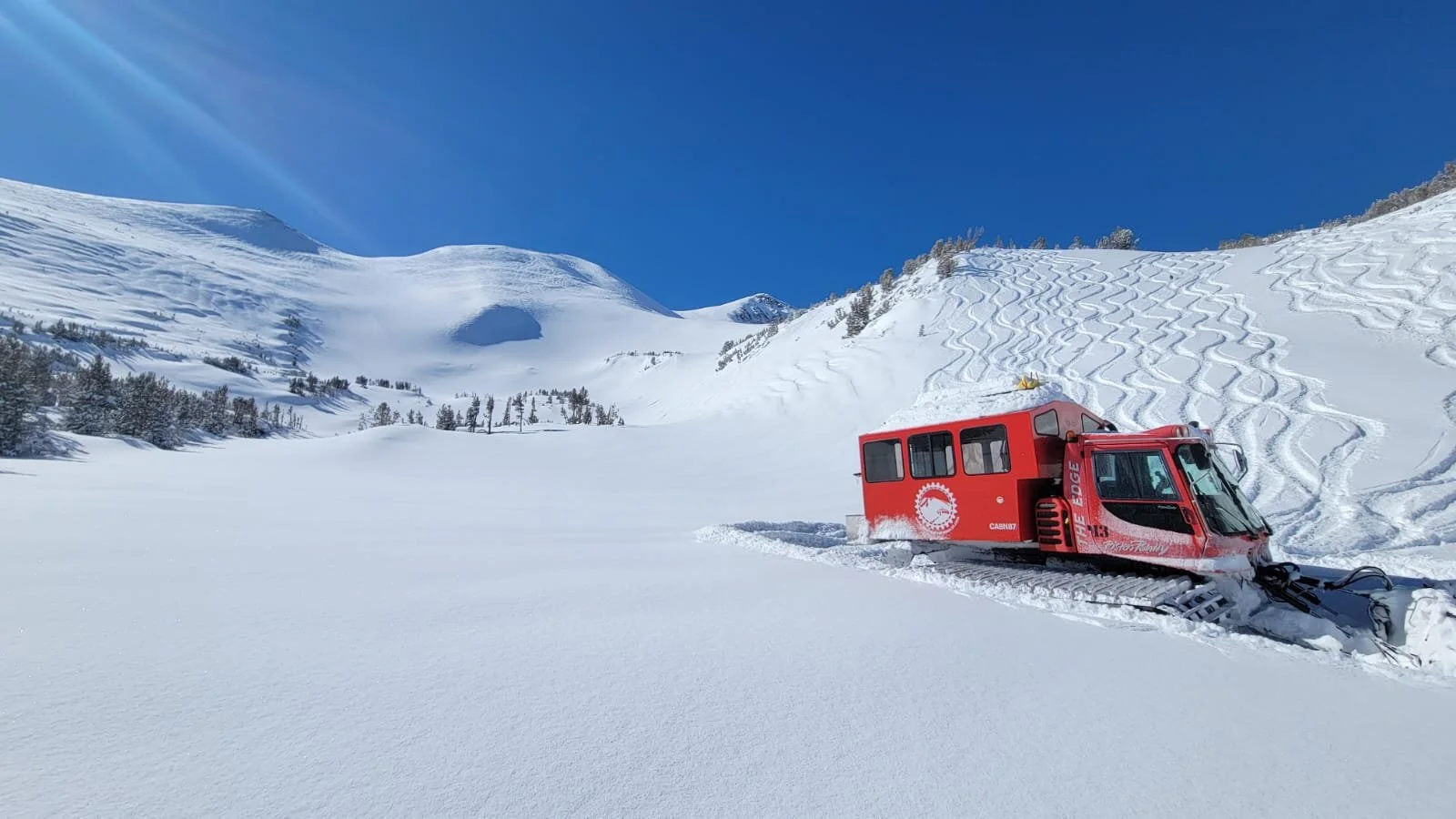 A red snowcat vehicle on a snow-covered mountain slope under a clear blue sky, with tracks in the snow and sparse trees on the hillside.