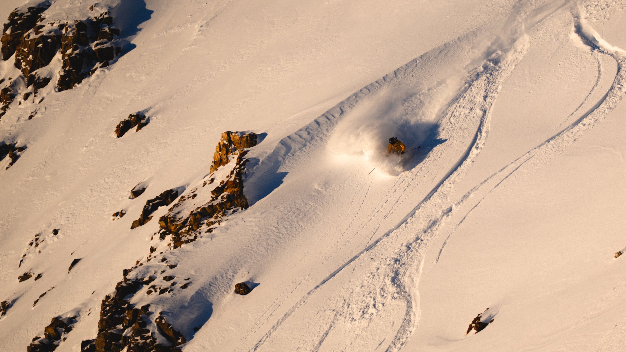 A skier descending a snowy mountain slope with rocks on one side, leaving trails in the snow.