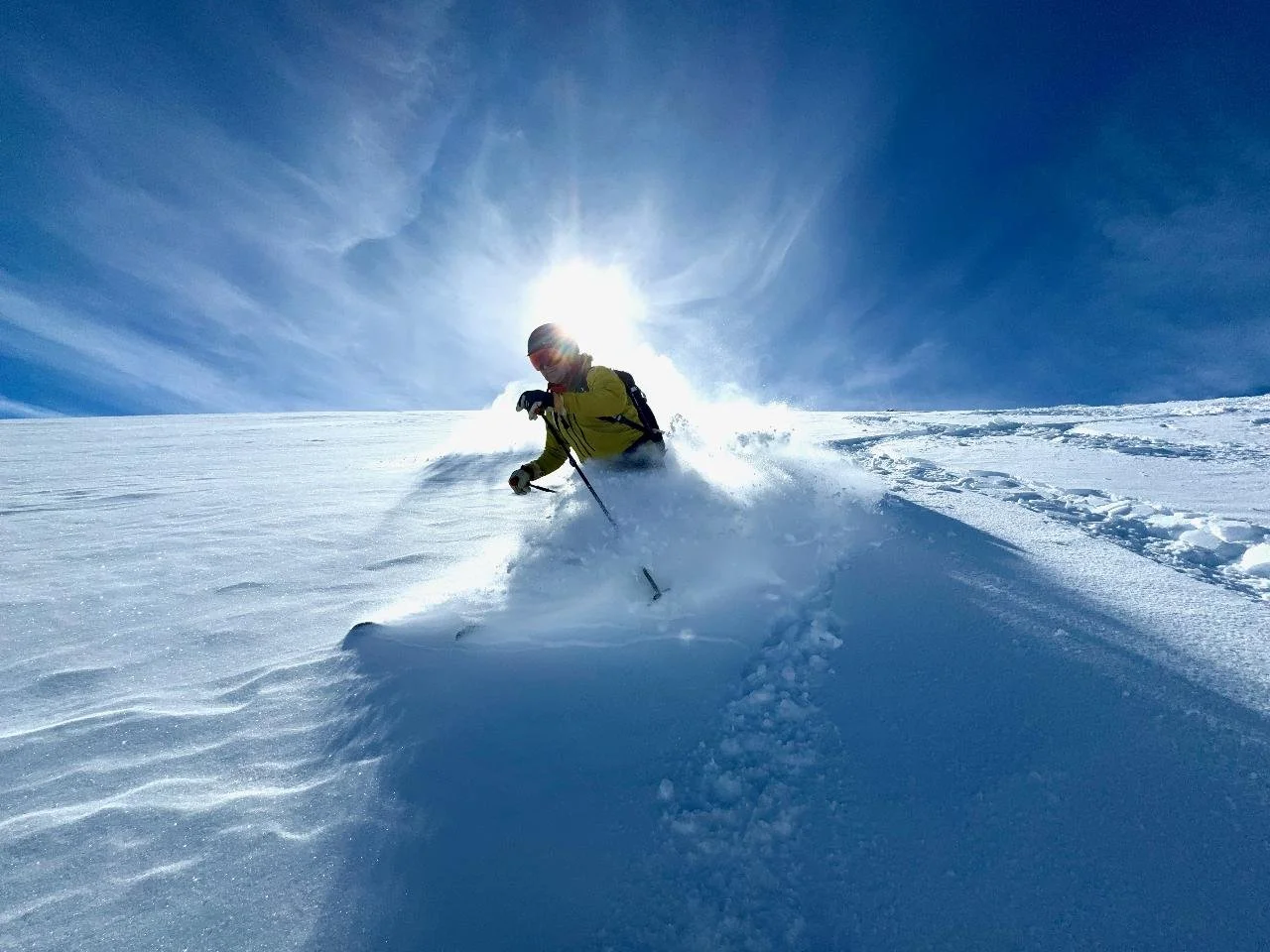 A person skiing downhill on snow covered mountain under a bright blue sky with the sun behind them.