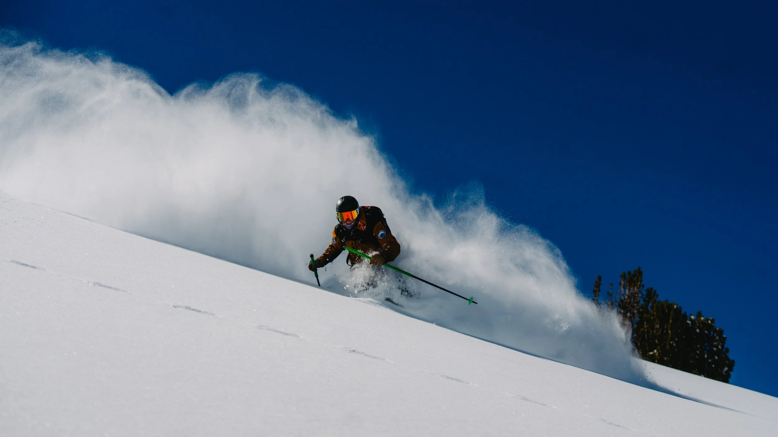 A person skiing down a snowy mountain slope, wearing a brown jacket, black helmet, orange goggles, and holding green ski poles, with snow spray and a clear blue sky.