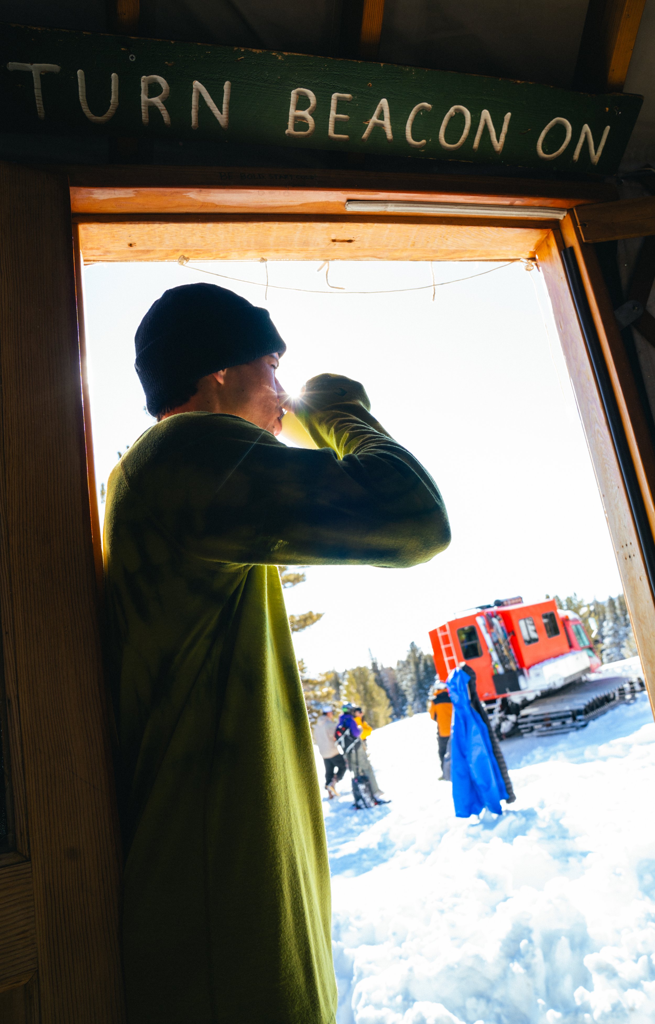 A person wearing a black beanie and green jacket is standing in a wooden structure, drinking from a yellow cup. Outside, on a snowy landscape, there are skiers and an orange snowcat vehicle.