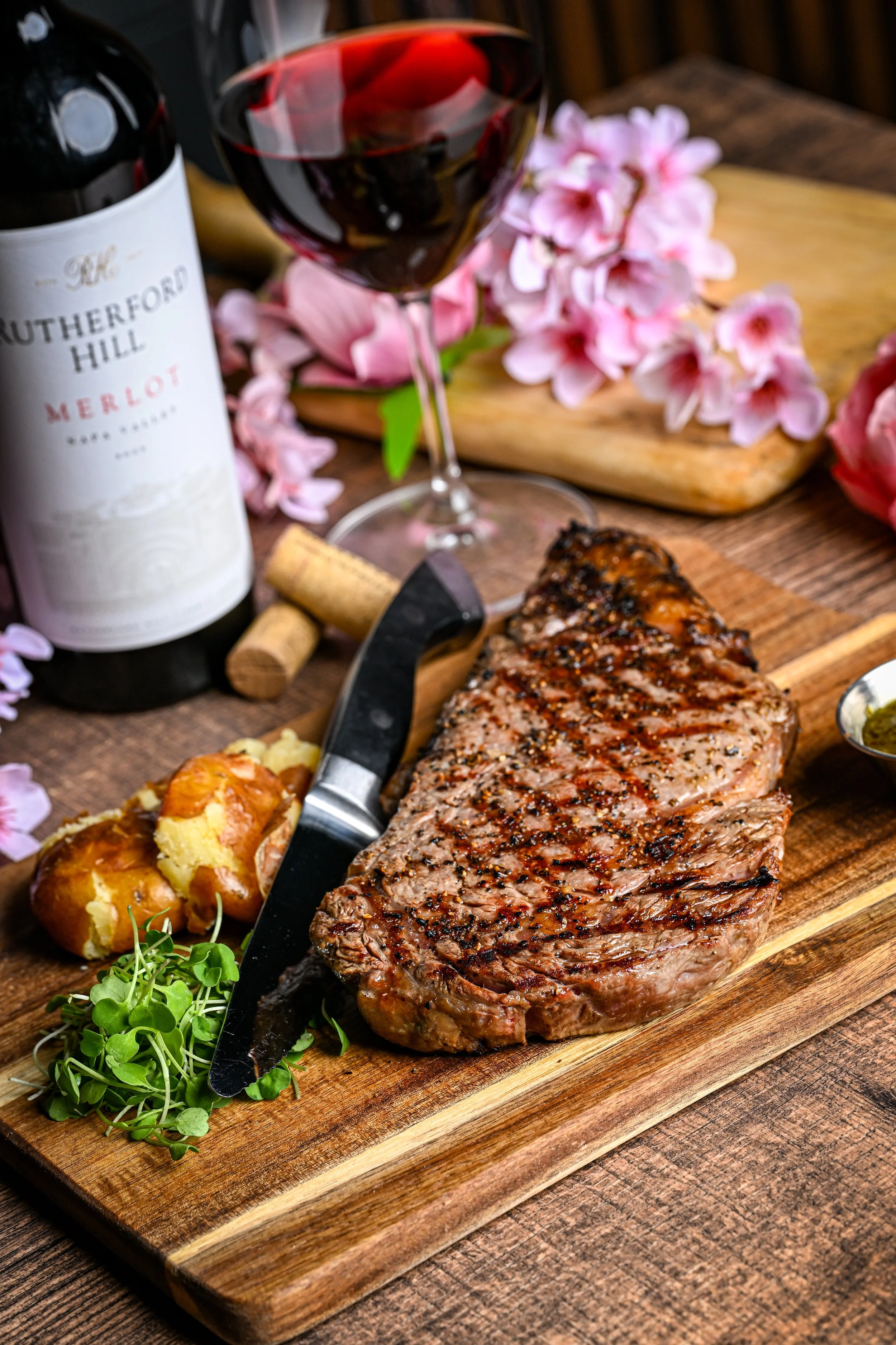 A wooden cutting board with a grilled steak, baked potato with toppings, microgreens, pink flowers, a glass of red wine, wine corks, a wine bottle, and a small dish of sauce.