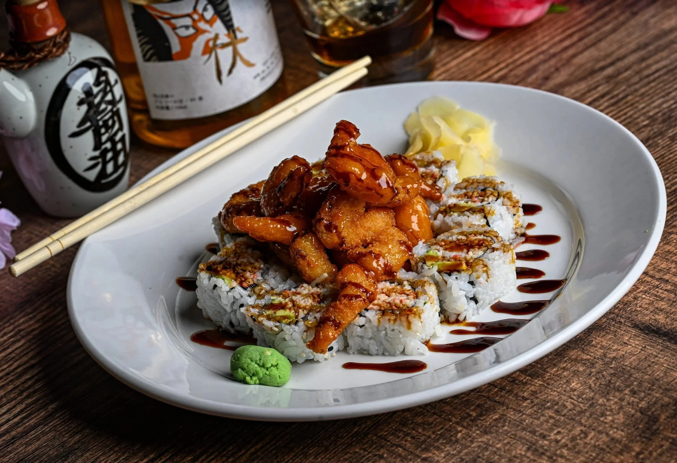 A white plate of sushi rolls topped with spicy orange sauce, with a small mound of wasabi on the side, garnished with drizzled sauce.