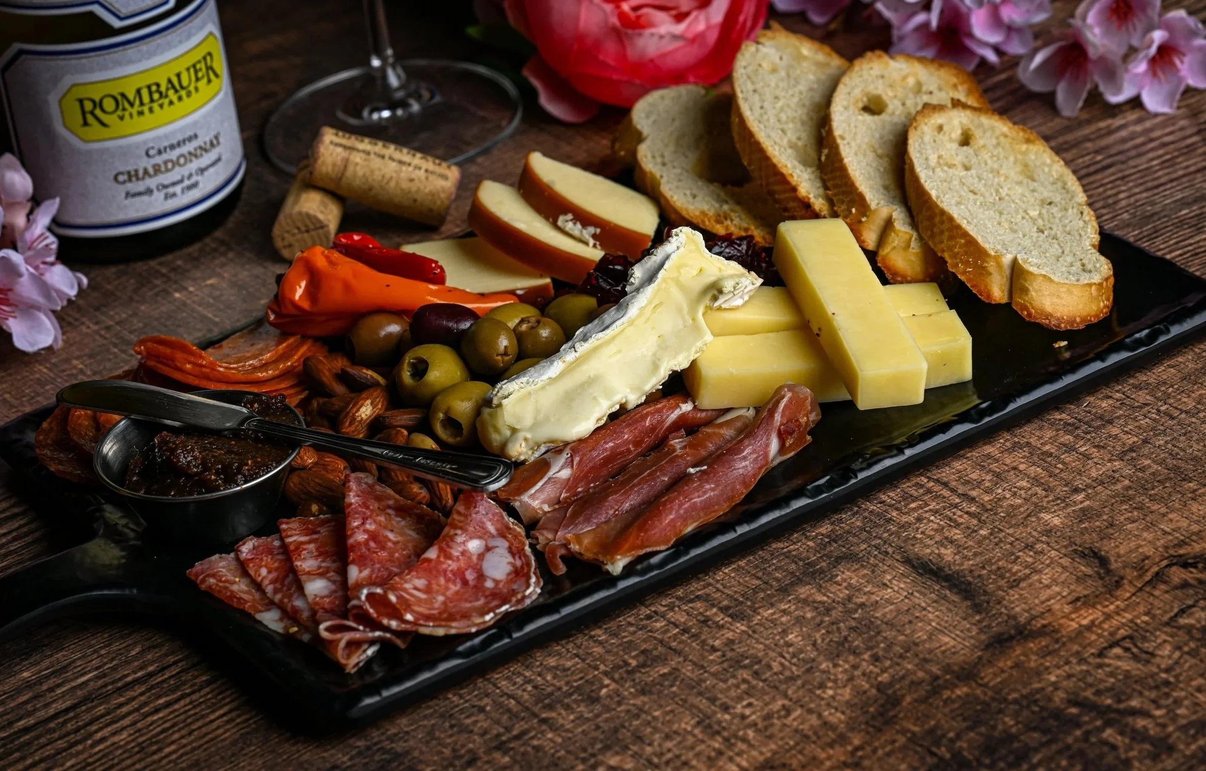 Charcuterie platter with sliced bread, cheeses, cured meats, olives, peppers, dried fruits, nuts, jam, and a bottle of wine on a wooden table, surrounded by pink flowers.