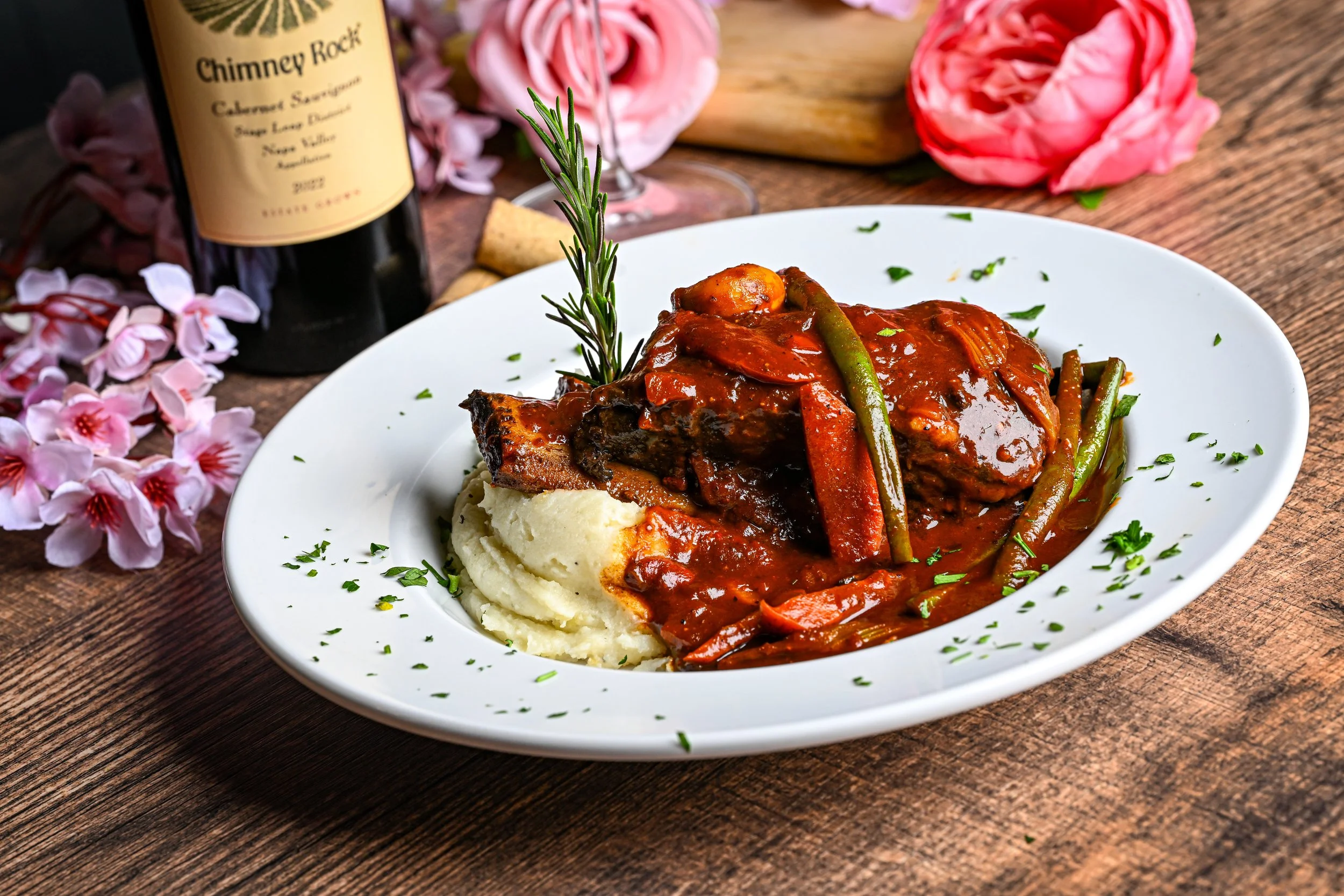 A plate of braised beef with vegetables and mashed potatoes, garnished with chopped herbs, on a wooden table with pink flowers and a bottle of Chimney Rock Cabernet Sauvignon in the background.