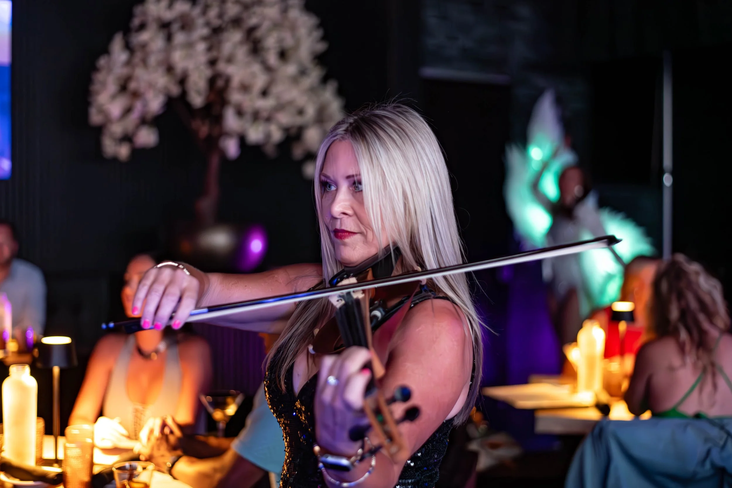 A woman with blonde hair playing a violin at a lively bar or restaurant with dim lighting, surrounded by people and colorful decorations.
