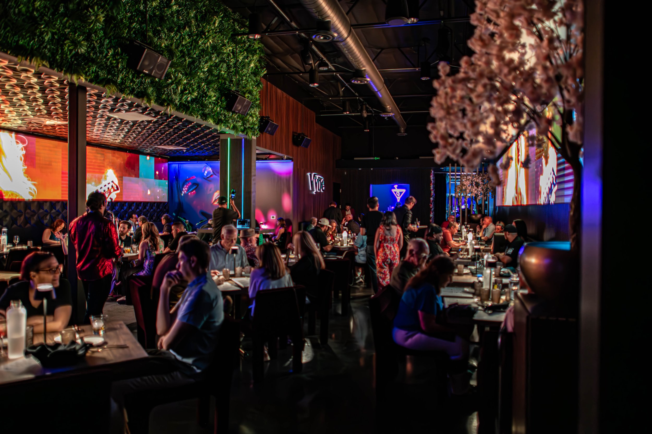 A busy indoor restaurant with many diners sitting at tables, colorful neon and digital screens on the walls, and pink cherry blossom decorations hanging from the ceiling.