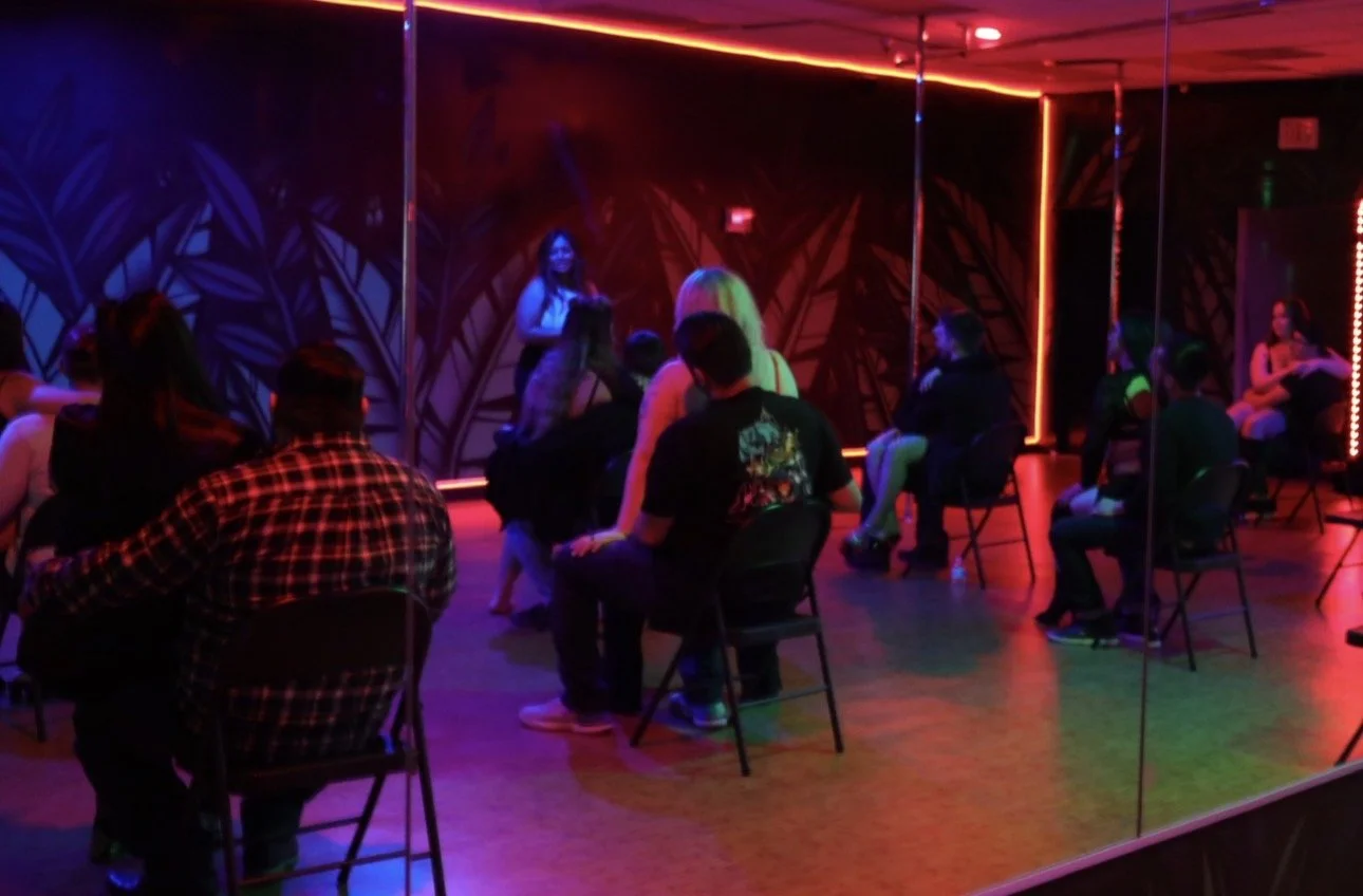 A group of people seated in a circle inside a dimly lit dance studio or club with neon red and blue lighting, a mirror wall studio space.  private lap dance lessons.  A way to get closer to your partner in a very intimate setting.