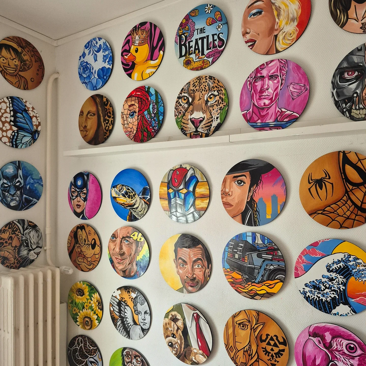 Une collection de tableaux colorés et variés accrochés sur un mur, représentant des personnages de dessins animés, des célébrités, des animaux et des icônes culturelles dans un style artistique diversifié.