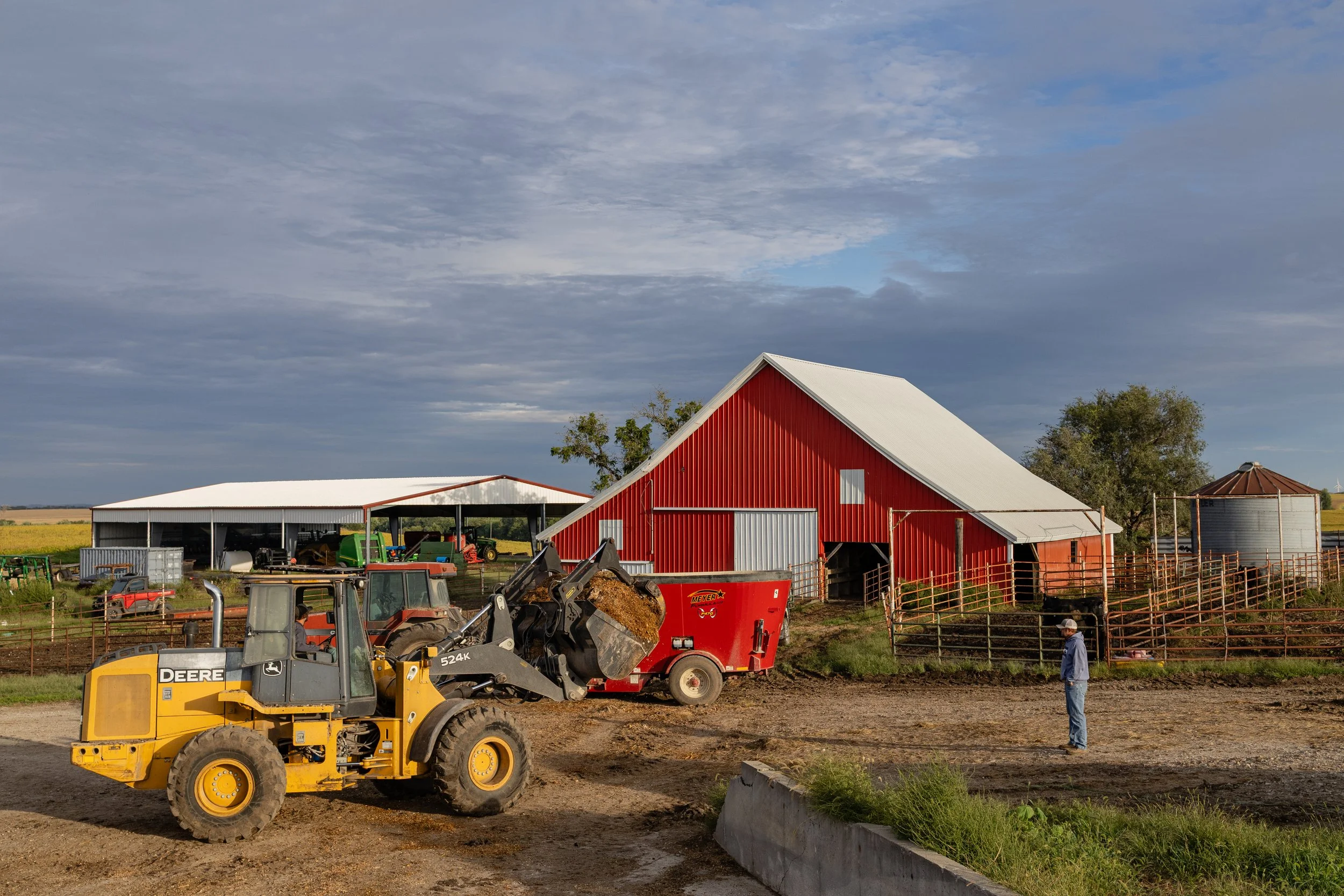 Russel, right, watches as his hired man Garrett Meyer, 26, uses an excavator to load corn silage into a large mixer with soybean meal to create a high energy–high protein feed on his farm near Maryville. The red barn in the background was built by fi