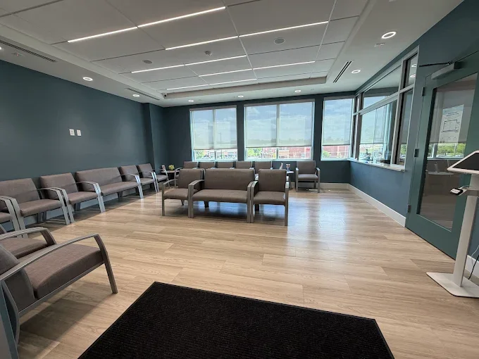 Empty waiting room with chairs along the walls, large windows, and a black mat on wooden floor.