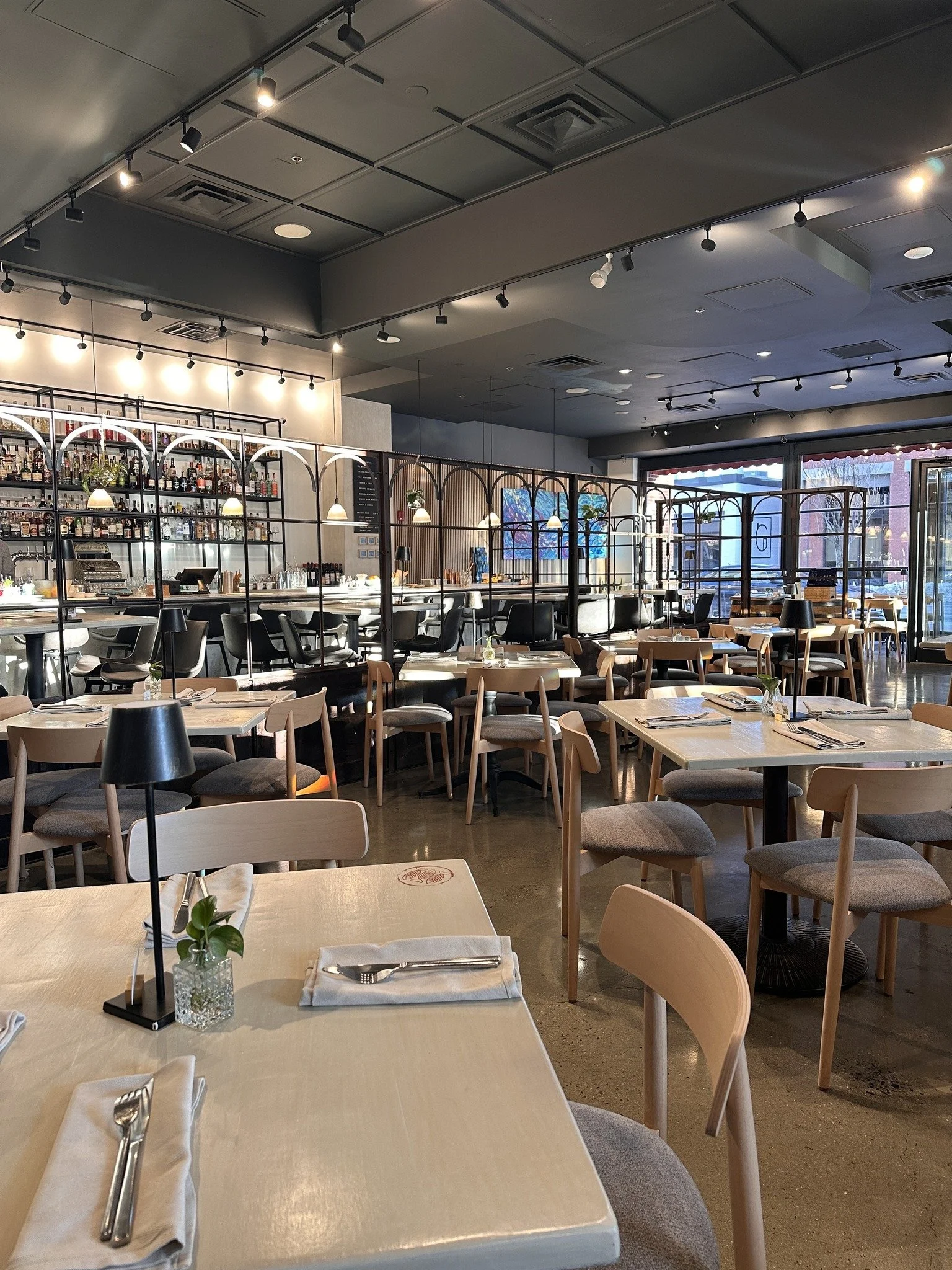 Interior of a modern restaurant with light wood tables and chairs, a bar with multiple bottles of alcohol, and decorative lighting fixtures. Large windows let in natural light, and artwork decorates the wall.