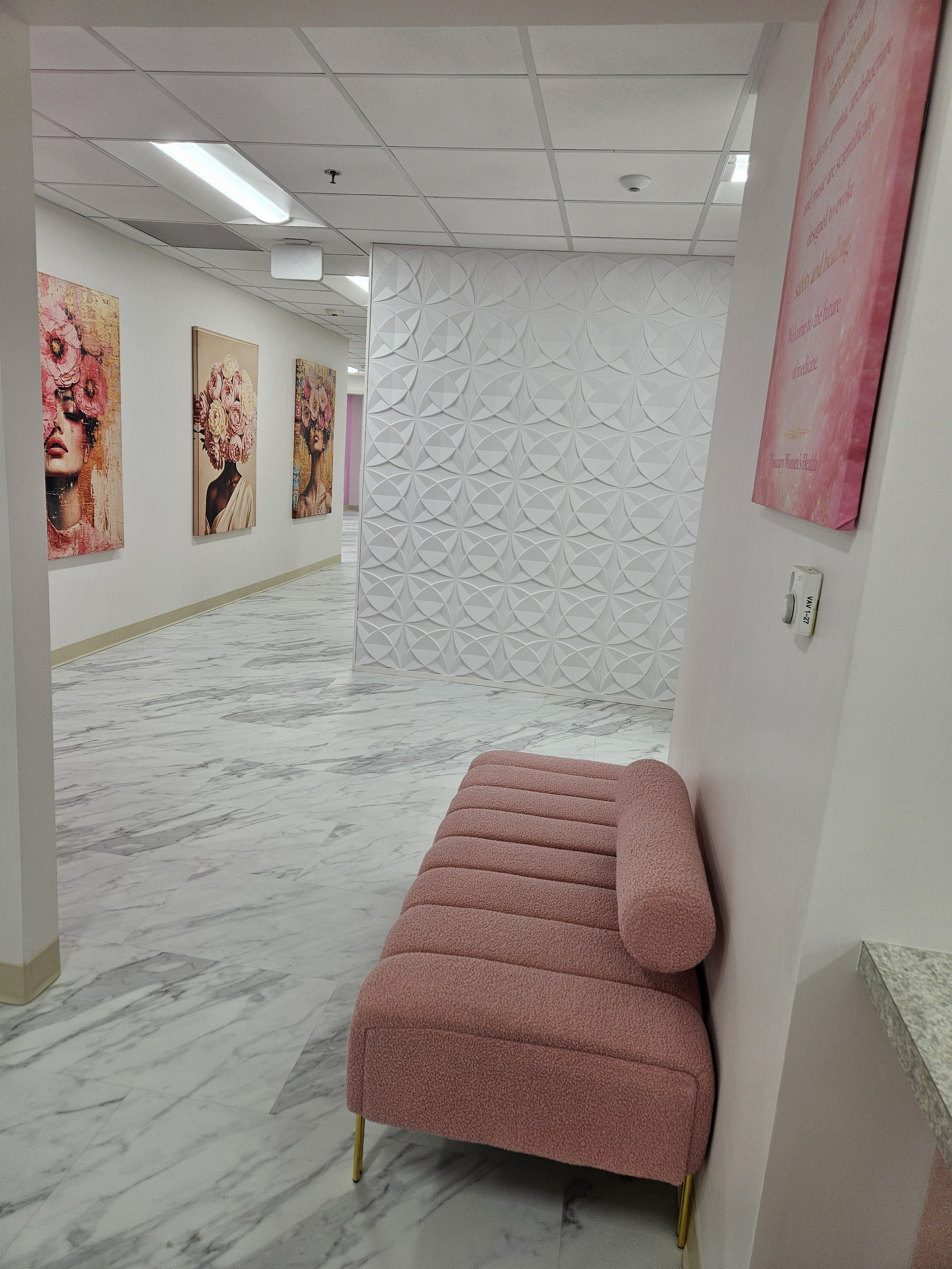 A waiting area with a pink upholstered bench and a white textured wall. Artwork of women with floral headpieces decorates the adjacent wall. The floor is marble, and the ceiling has fluorescent lighting.