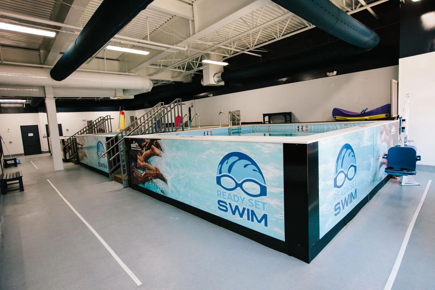 Indoor swimming pool with a floating platform marked with the ‘Ready Set Swim’ logo, water safety signs, colorful rescue equipment, and benches around the pool area.