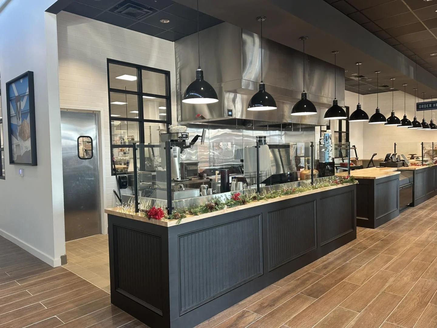 Indoor view of a modern foodservice counter with black cabinets, black lighting fixtures, and a glass sneeze guard. The area features a coffee machine and beverage dispensers, with a decorated counter featuring greenery and red berries, in a well-lit space with wood flooring.