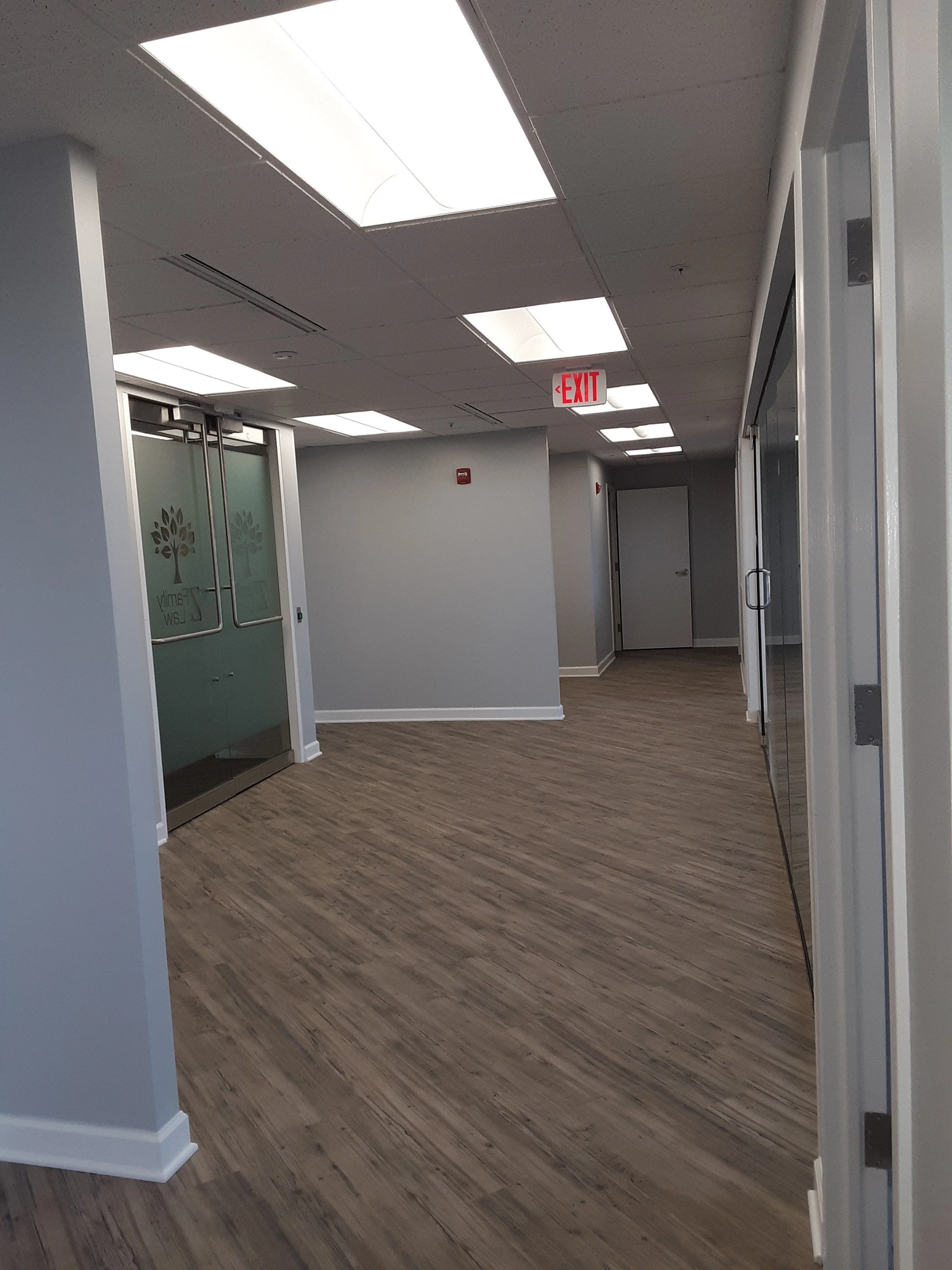 An empty office hallway with wood flooring, gray walls, ceiling lights, glass doors, and an exit sign hanging from the ceiling.