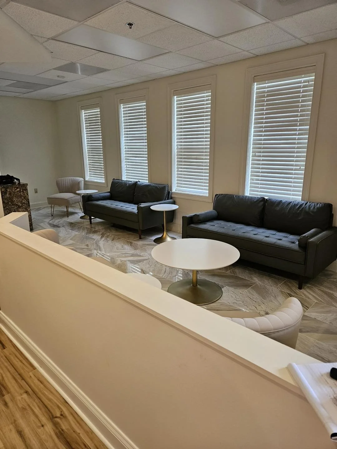A modern waiting room with two black sofas, a white armchair, three small round tables, and four large windows with blinds, beige walls, and a tiled floor.