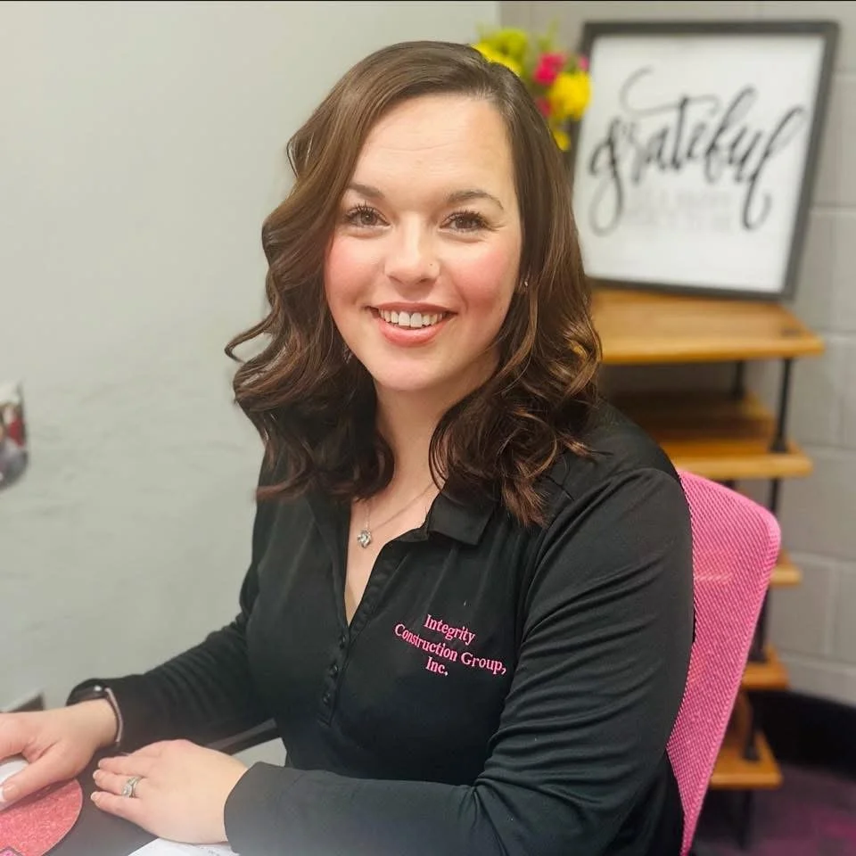 A smiling woman with shoulder-length curly brown hair sitting at a desk in an office. She is wearing a black polo shirt with pink embroidery and a silver necklace. There is a pink chair, a flower arrangement, and a framed sign that reads 'grateful' in the background.